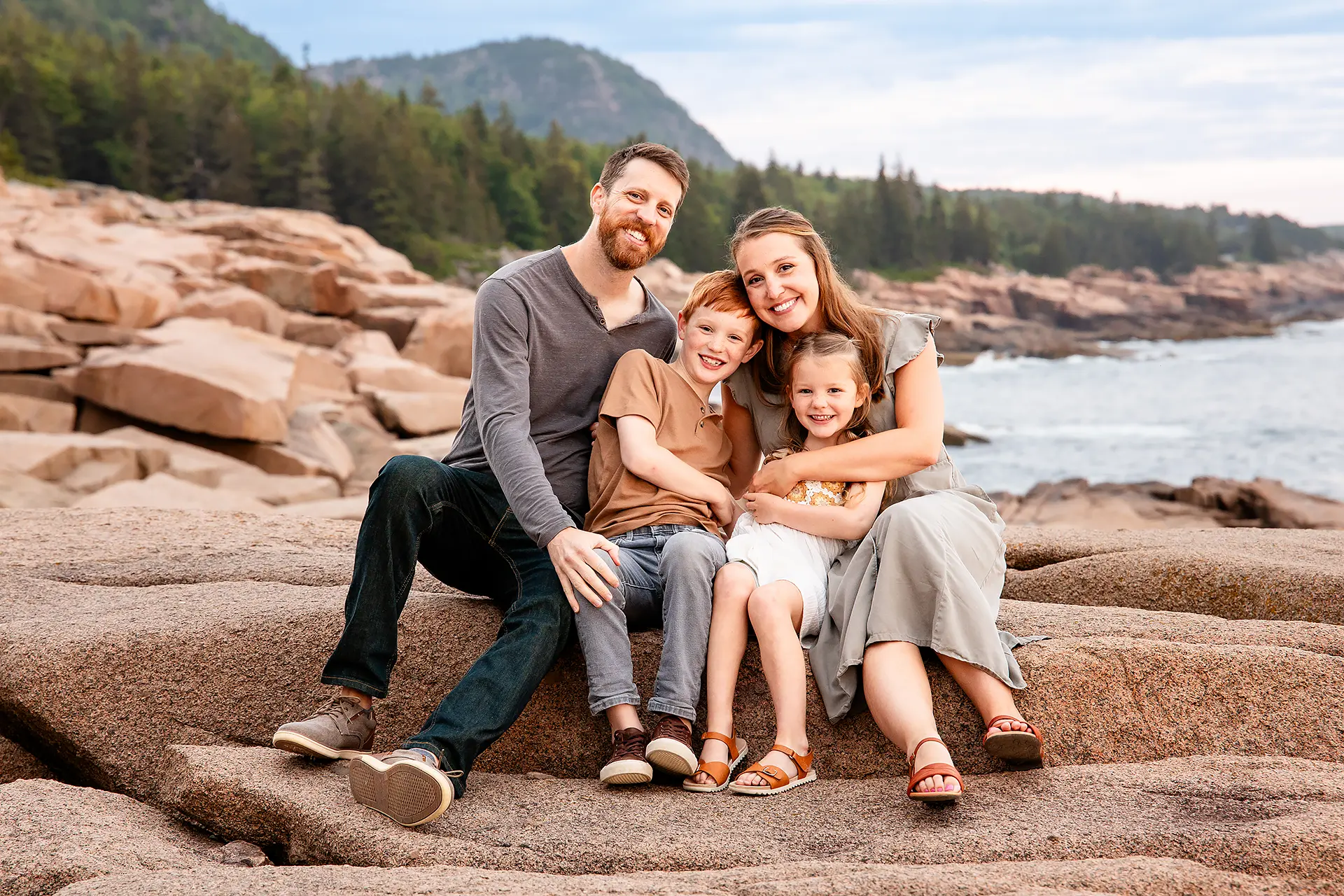 Two young parents sit with their children on a cliff near the ocean in Acadia National Park outside of Bar Harbor, Maine.
