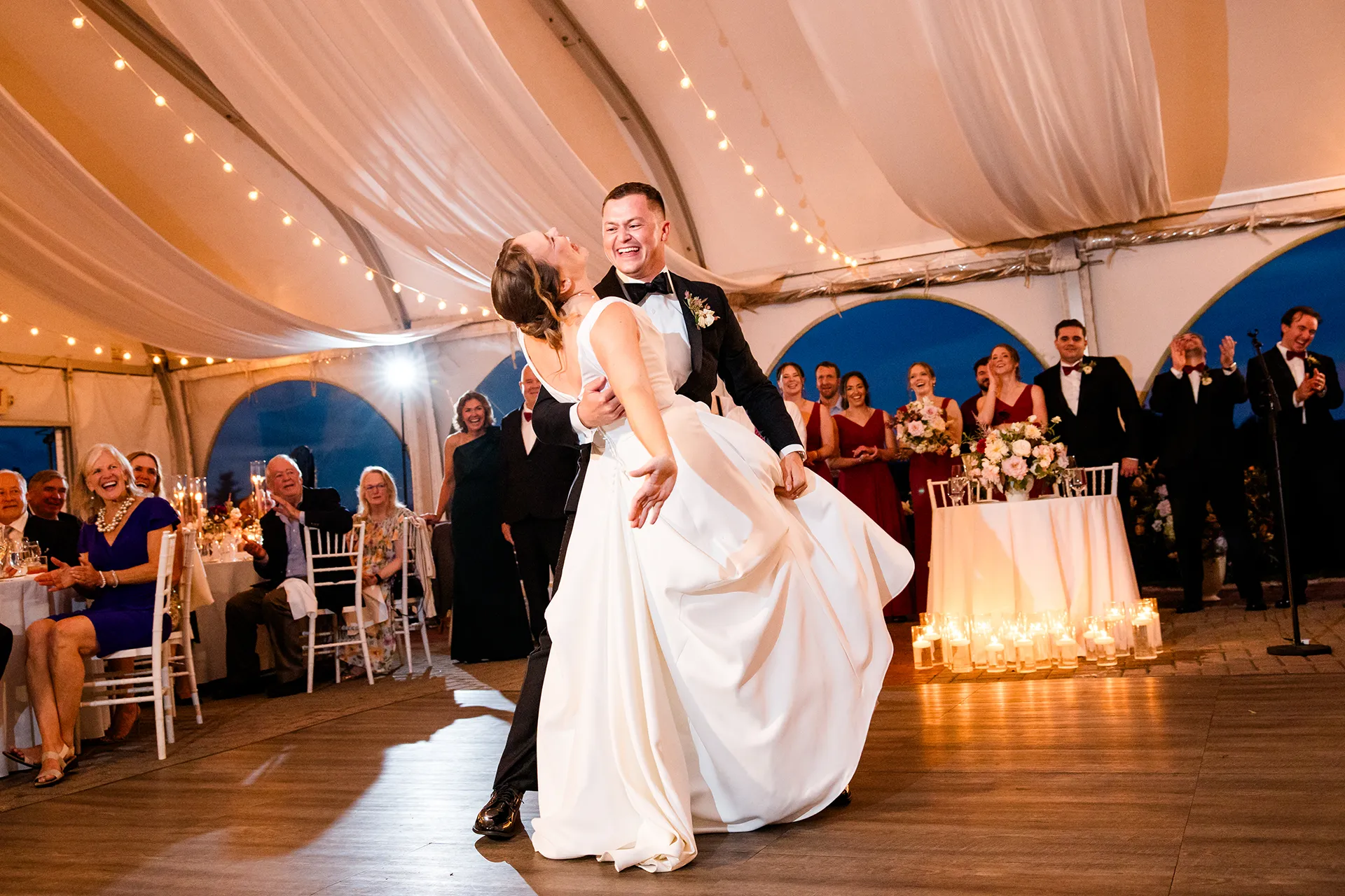 During a first dance a groom dips a bride while they laugh as guests celebrate behind them during a tented wedding reception in Maine.