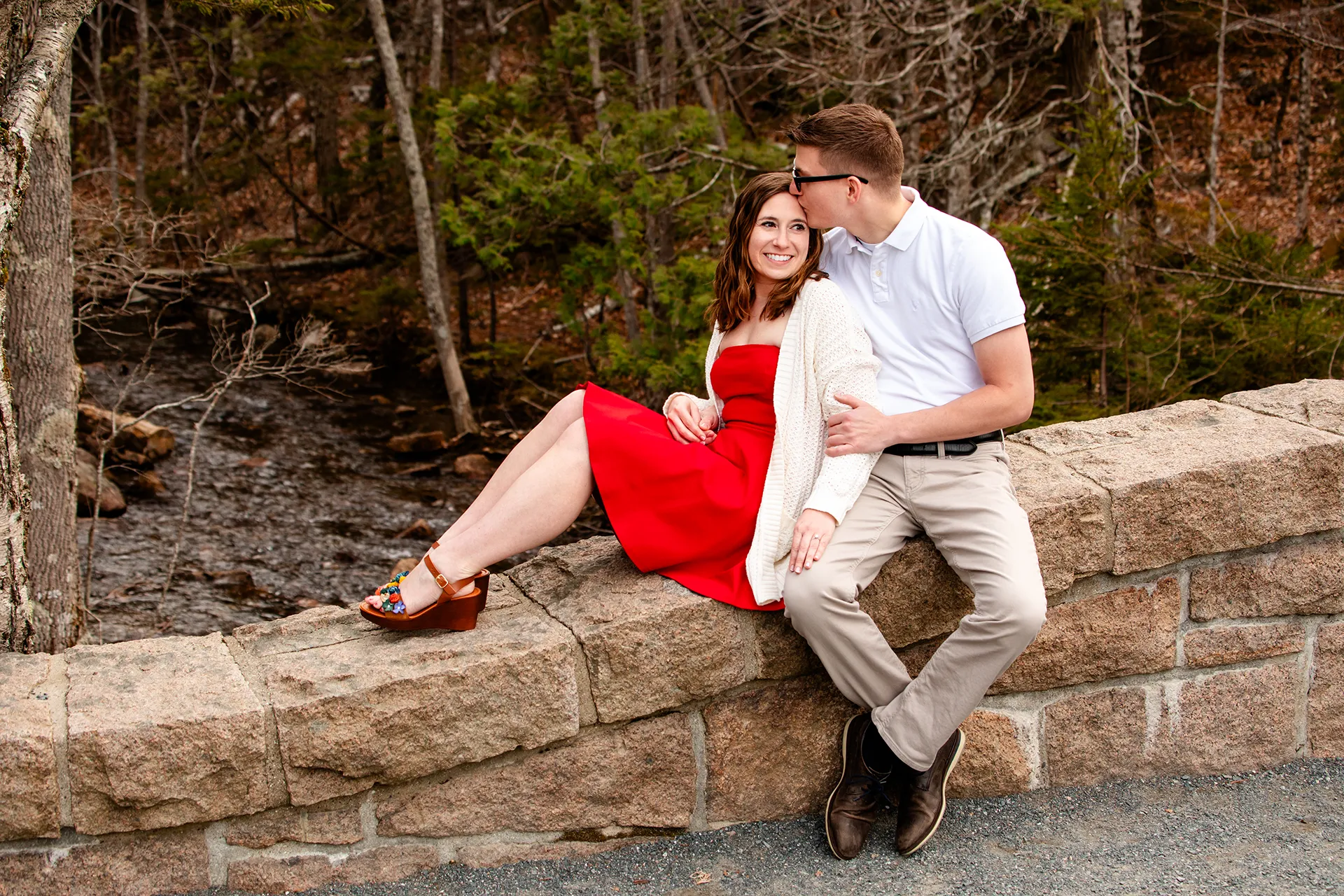 maine engagement photography chelsea mike 02 A man kisses a woman on the head as they sit on a stone bridge at Jordan Pond in Acadia National Park, Maine.