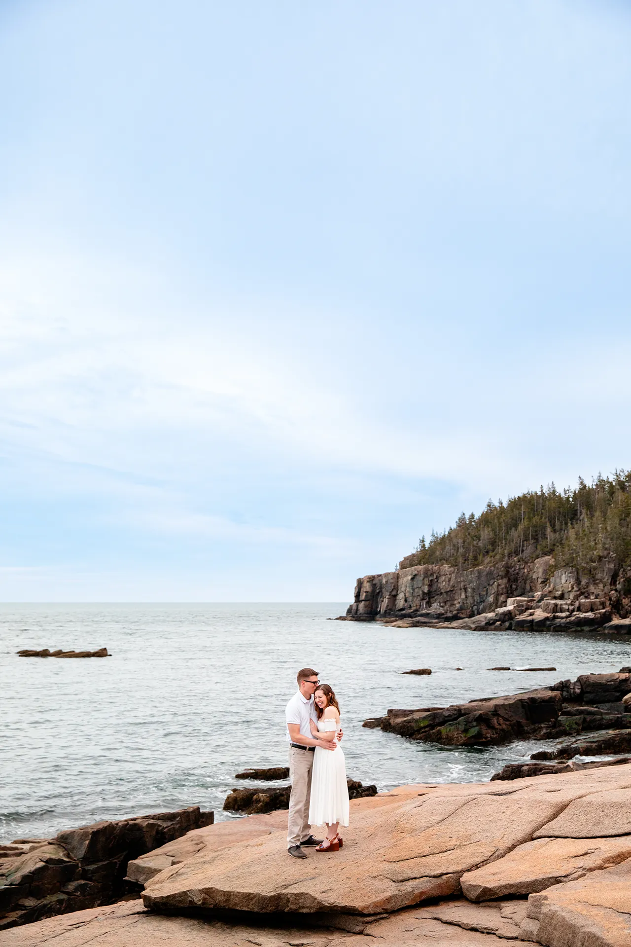 maine engagement photography chelsea mike 21 A woman laughs as a man kisses her on the head as they stand on a cliff overlooking the ocean in Acadia National Park, Maine.