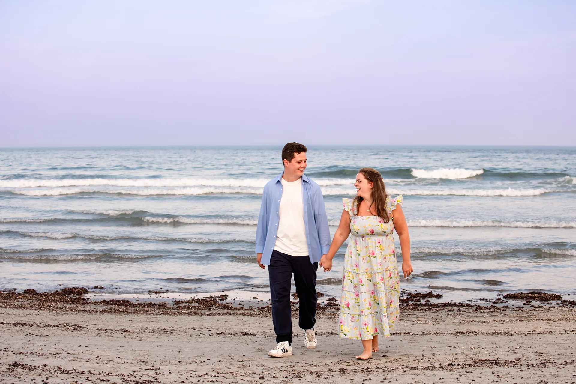 A man and woman hold hands and walk during engagement portraits on Long Sands Beach in York, Maine.