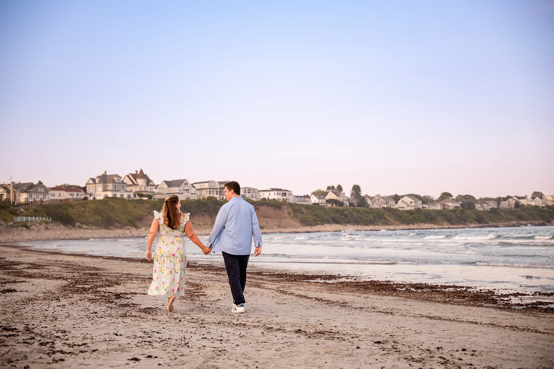 A newly engaged couple hold hands and walk on Long Sands Beach during engagement portraits in York, Maine.
