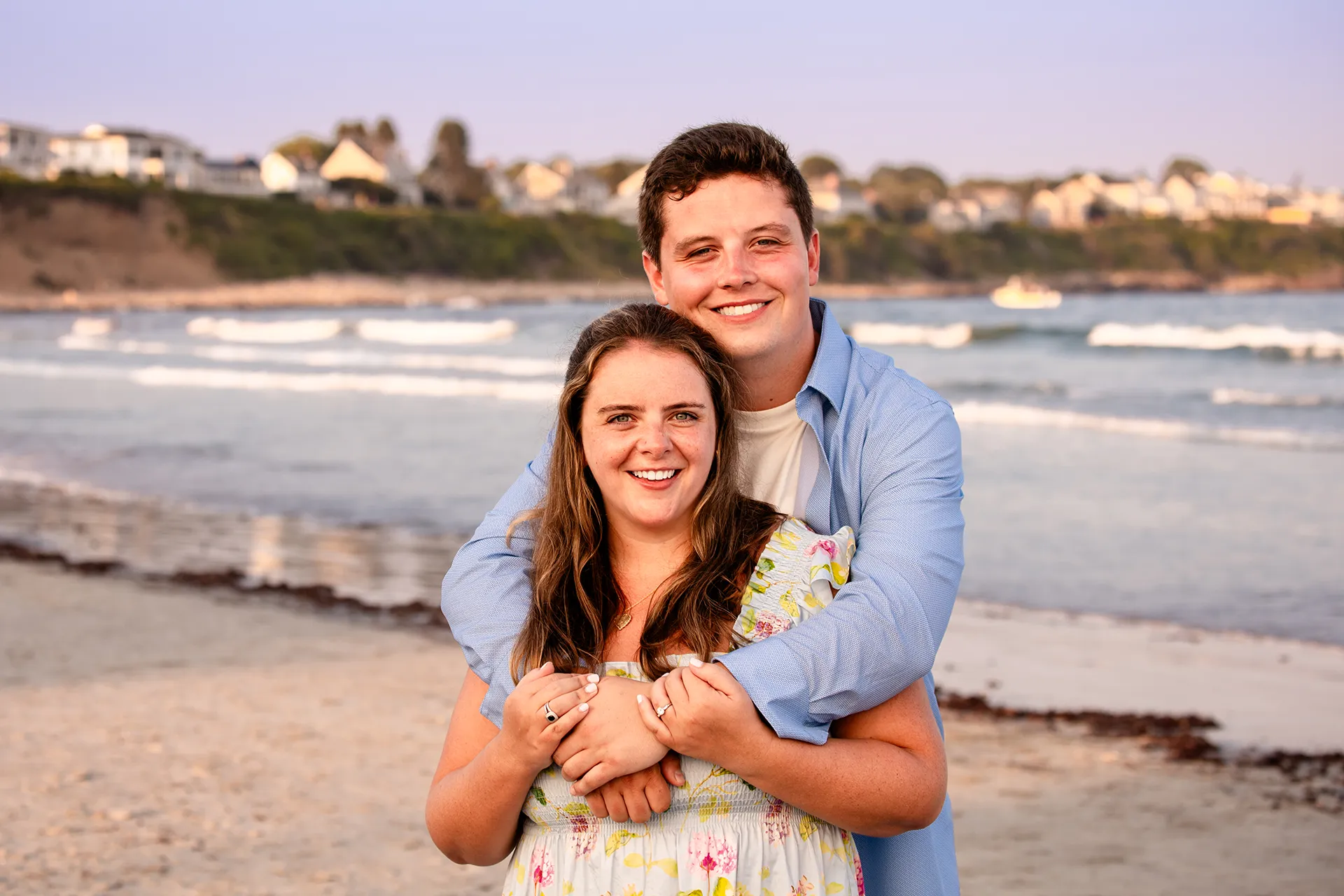 A man hugs a woman as they smile and pose during engagement portraits on Long Sands Beach in York, Maine.