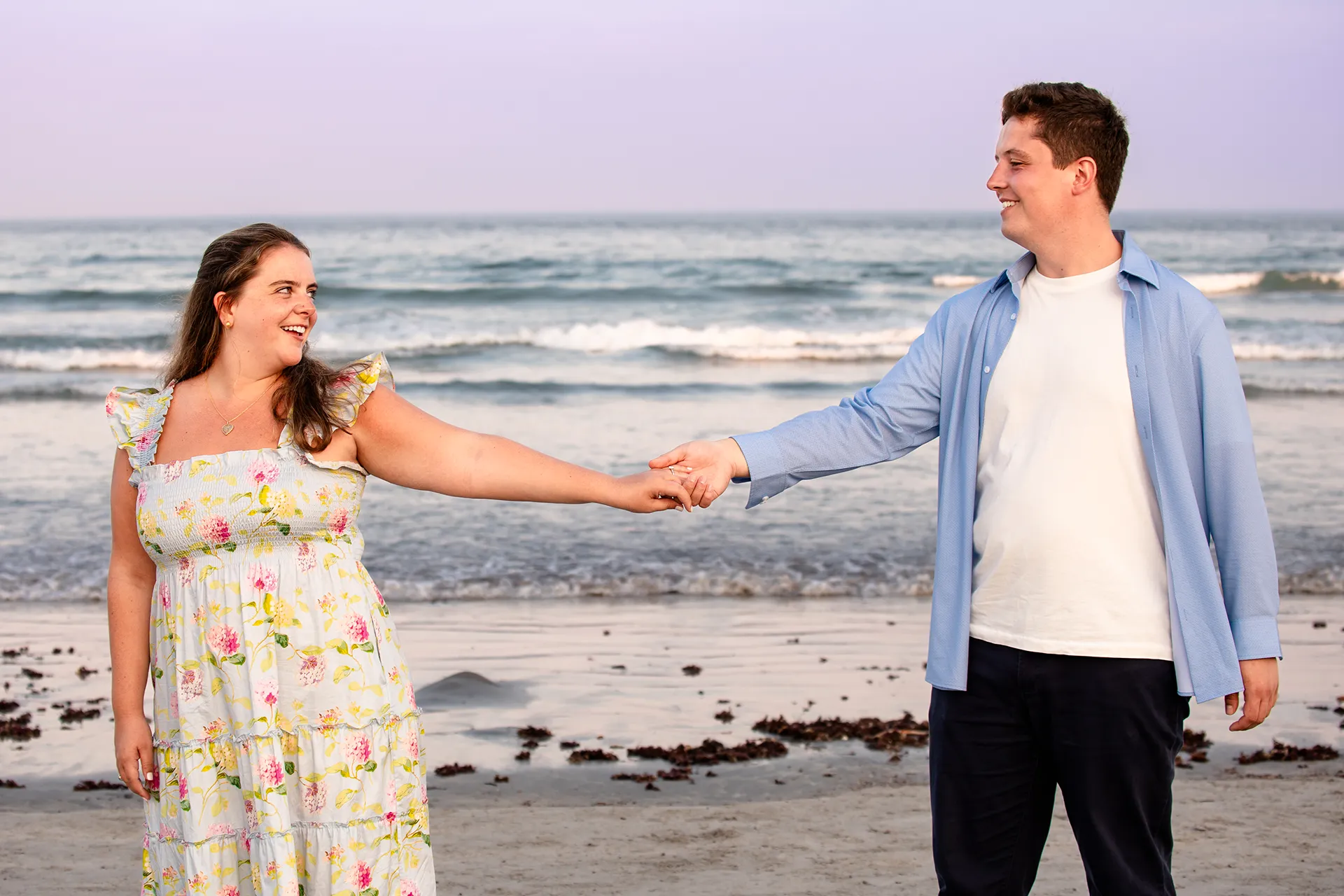 A man and woman hold hands and smile at each other during engagement portraits on Long Sands Beach in York, Maine.