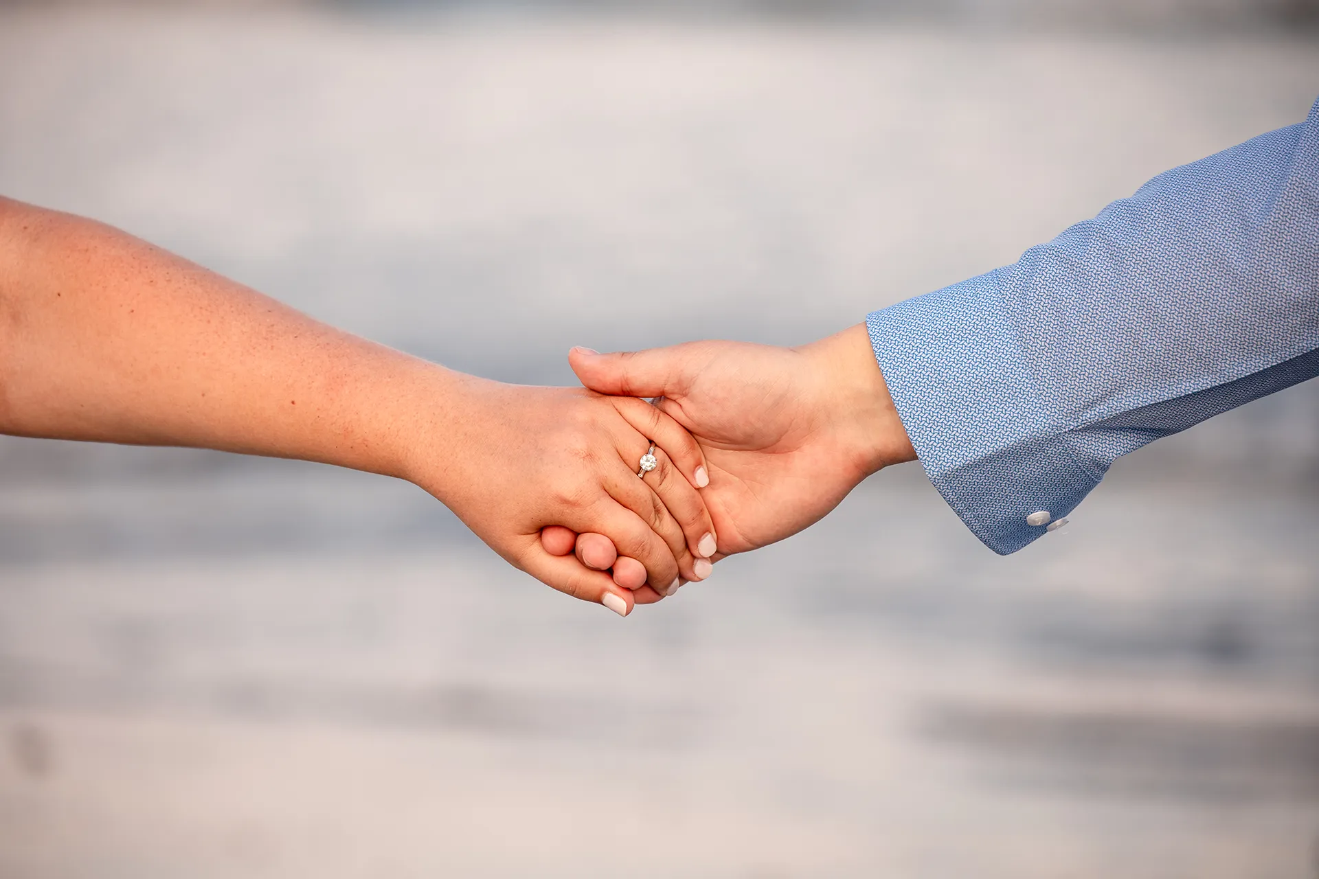 A closeup of a man's hands holding a woman's hand wearing an engagement ring on Long Sands Beach in York, Maine.
