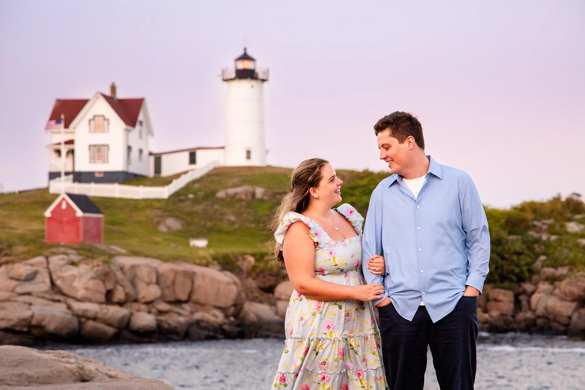 A newly engaged couple smile at each other during sunset engagement portraits at Nubble Lighthouse in York, Maine.