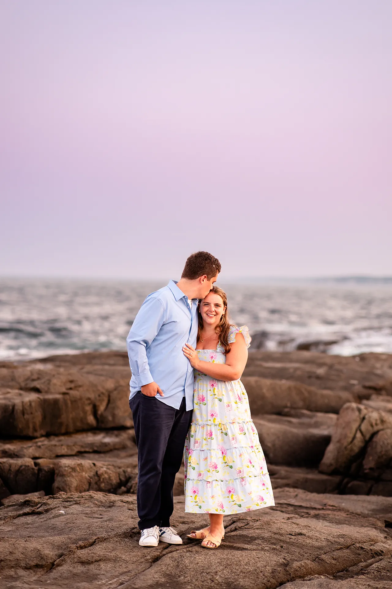 A man kisses a smiling woman while they pose at sunset during an engagement session at Nubble Lighthouse in York, Maine.