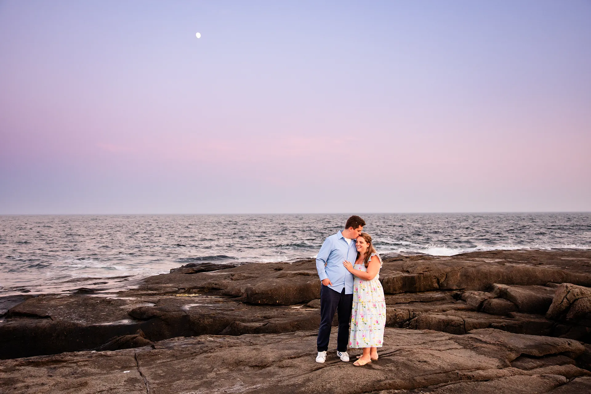 A woman smiles as a man kisses her on the head during sunset engagement portraits at Nubble Lighthouse in York, Maine.