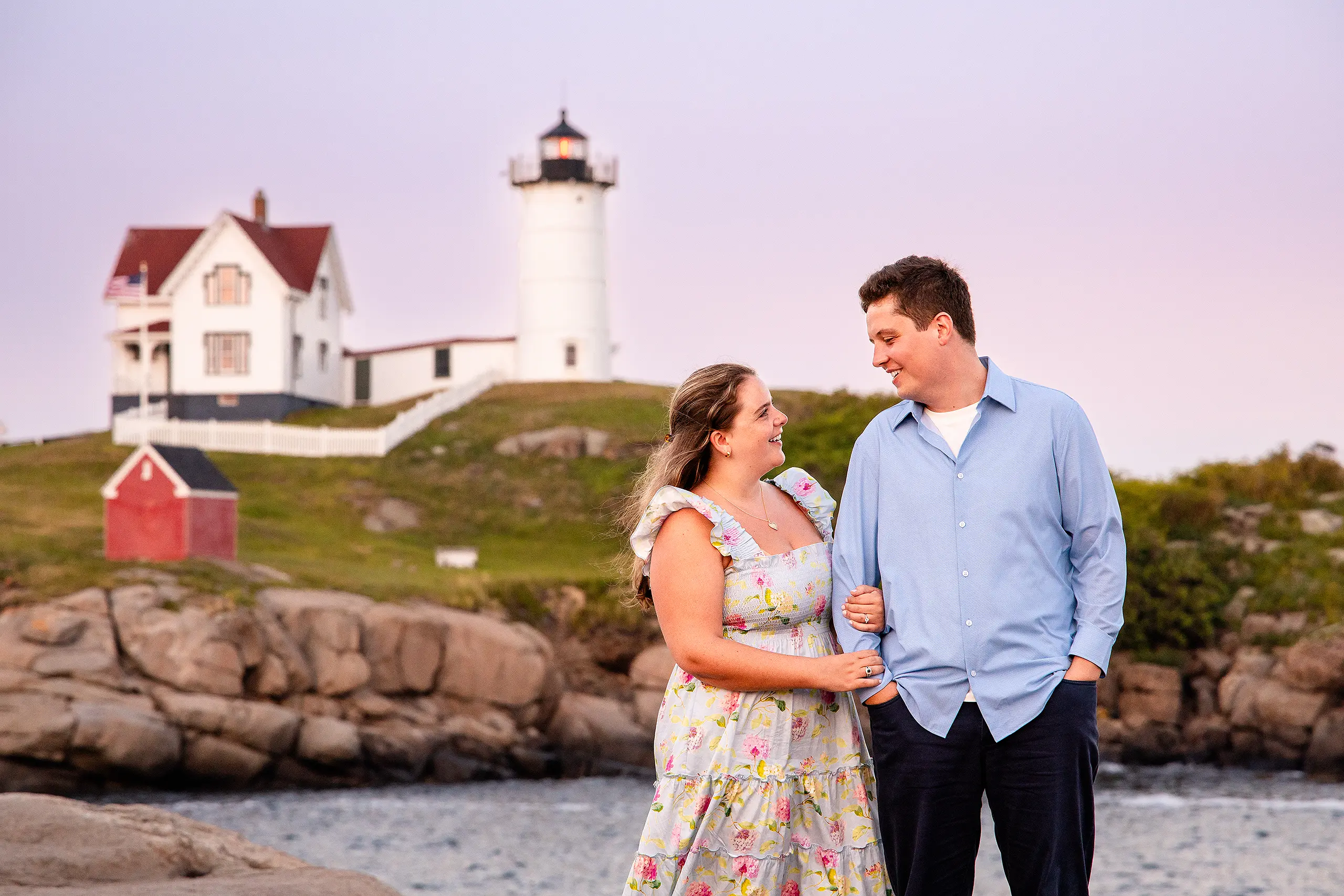 A newly engaged couple smile at each other during sunset engagement portraits at Nubble Lighthouse in York, Maine.
