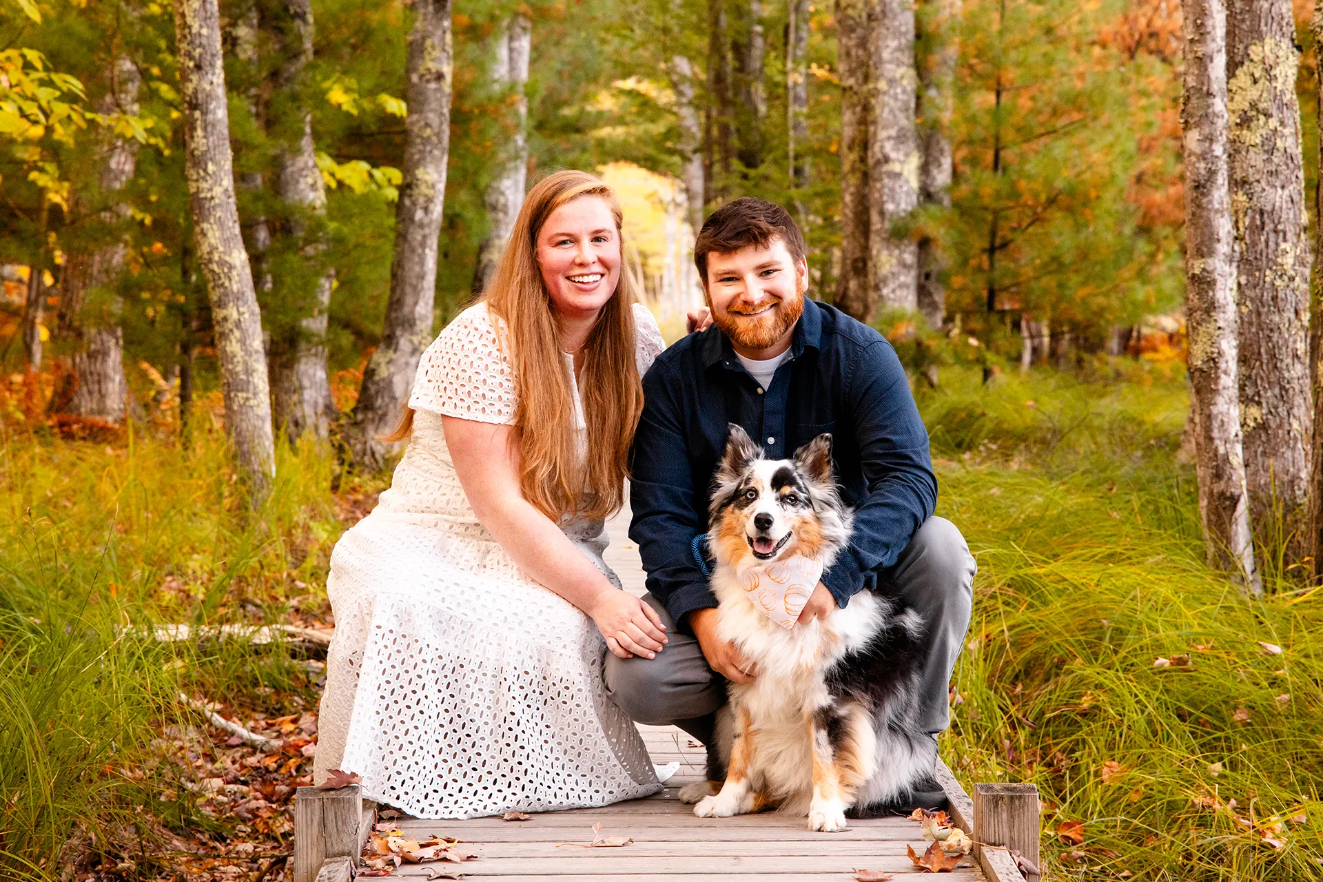 A man and woman smile and pose with their dog during an engagement session on the Jesup Path at Acadia National Park in Bar Harbor, Maine.