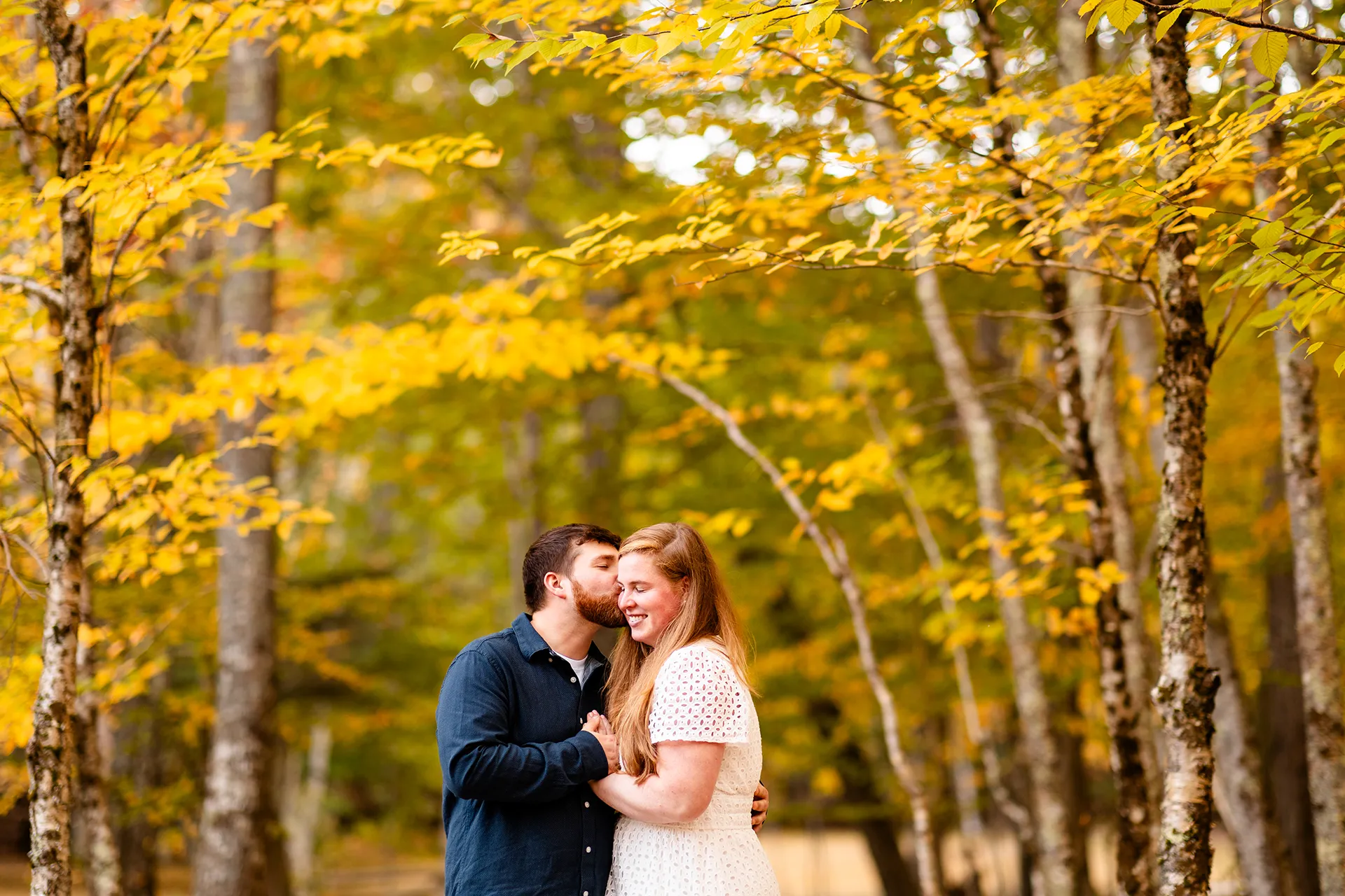 A woman smiles as a man kisses her on the cheek during a fall engagement session on the Hemlock Trail at Acadia National Park in Bar Harbor, Maine.