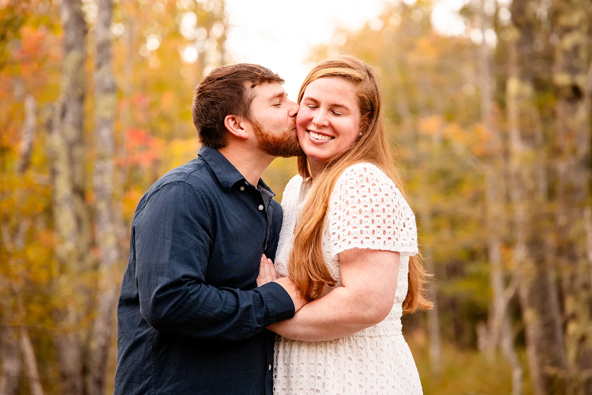 A man kisses a smiling woman on the cheek during a fall engagement session on the Jesup Path at Acadia National Park in Bar Harbor, Maine.