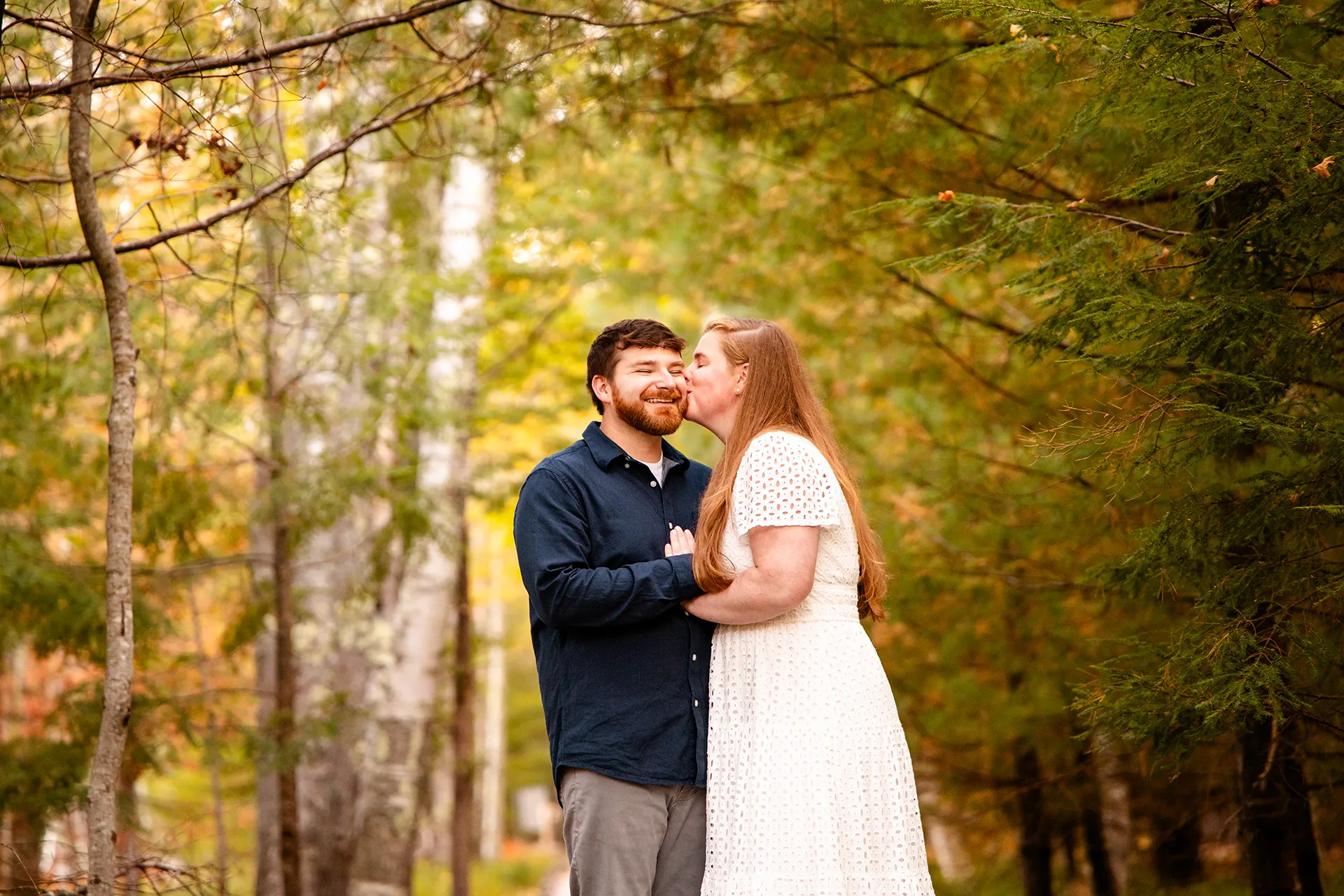 A man smiles as a woman kisses him on the cheek during an engagement session on the Jesup Path at Acadia National Park in Bar Harbor, Maine.