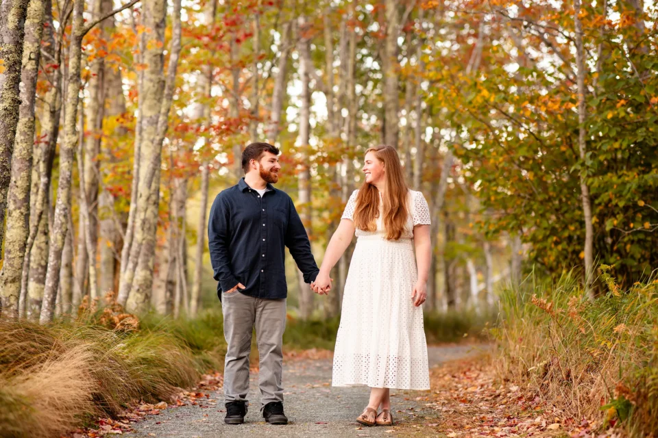 A man and woman hold hands and smile at each other during a fall engagement session at Acadia National Park in Bar Harbor, Maine.