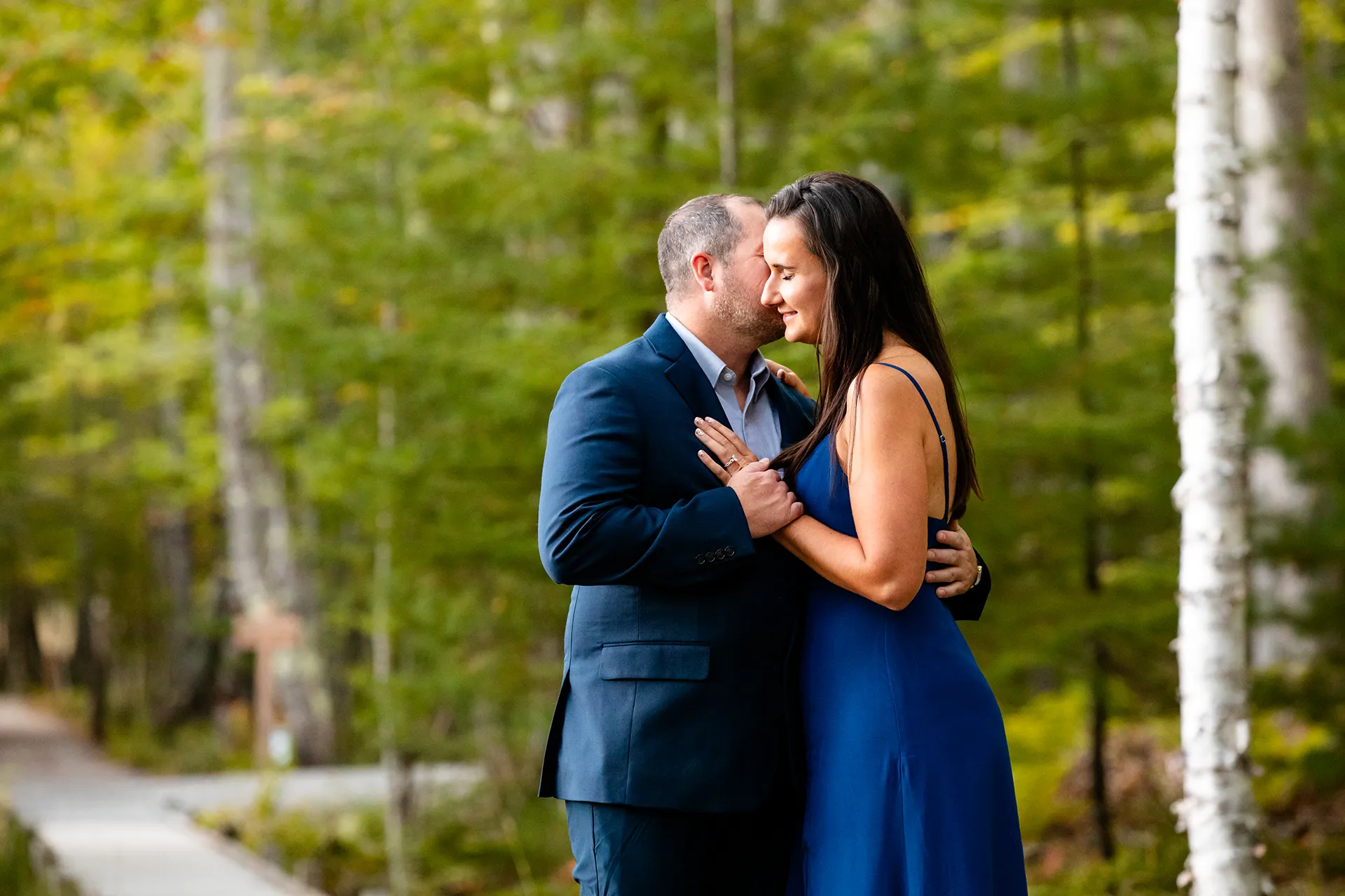 A woman smiles as a man kisses her on the cheek during a engagement session on the Jesup Path in Acadia National Park, Maine.
