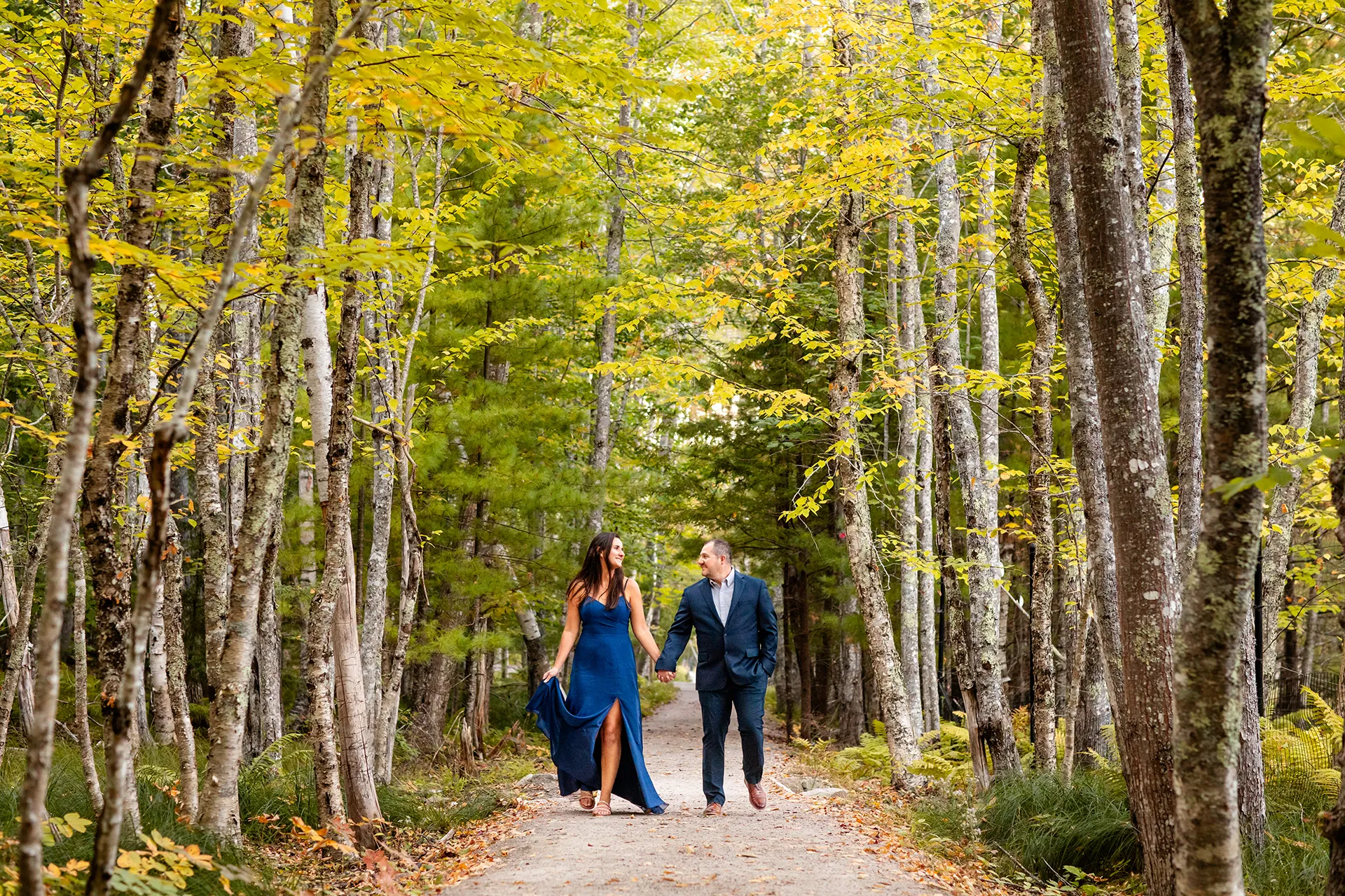 An engaged couple hold hands and walk during a engagement session on the Hemlock Trail near the Wild Gardens of Acadia in Acadia National Park.