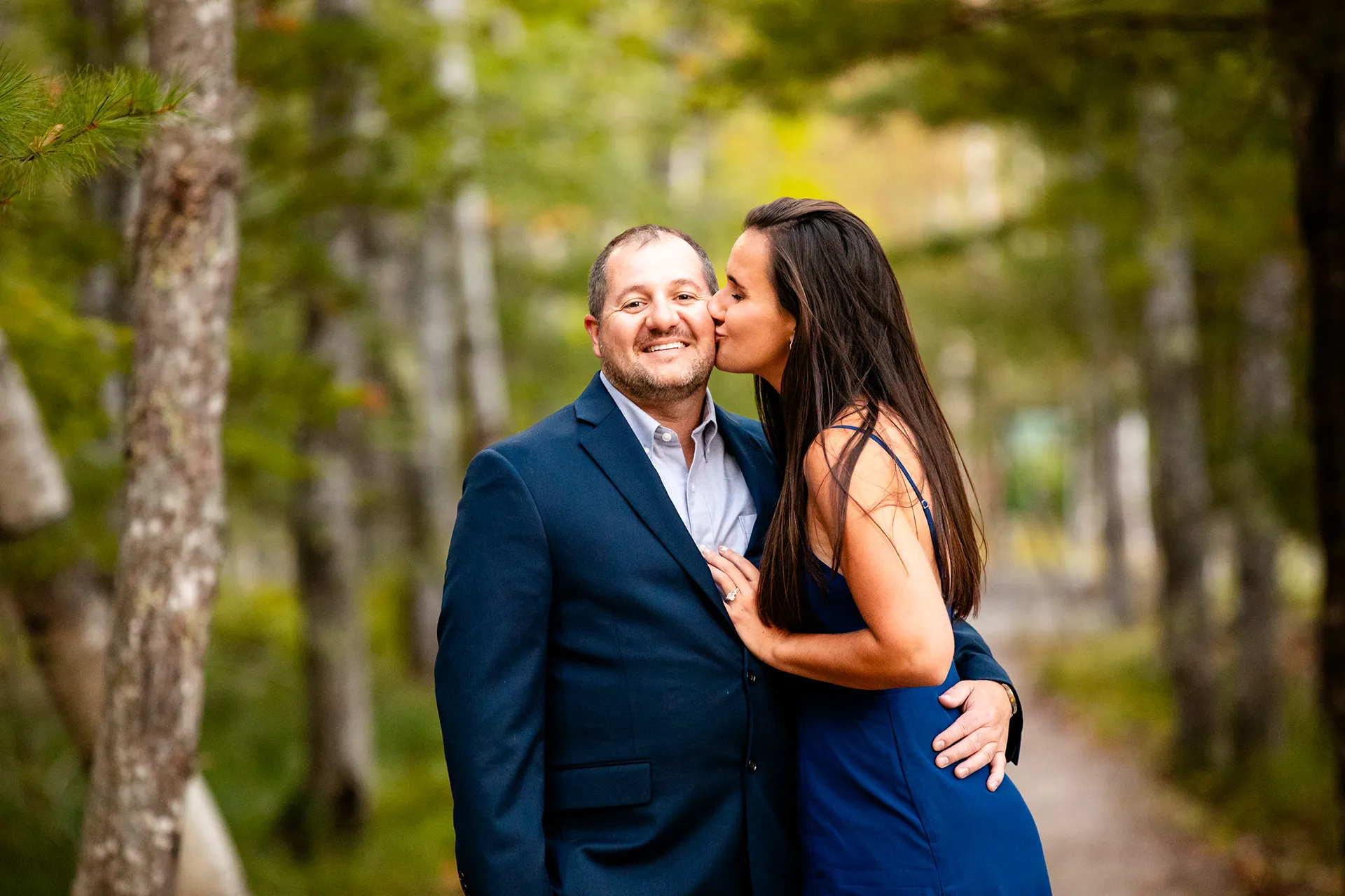 A man smiles as a woman kisses him a on the cheek during a engagement session on the Hemlock Path in Acadia National Park, Maine.