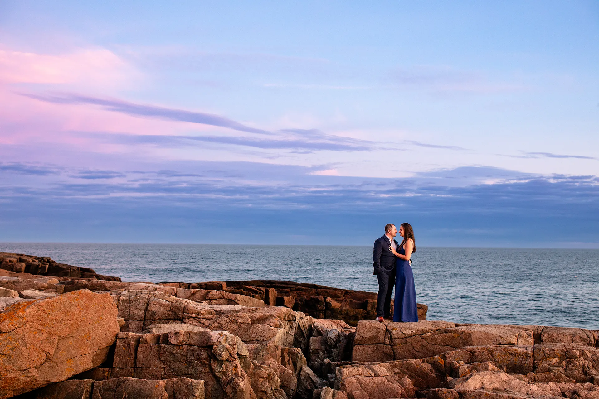 An engaged couple pose on a cliff as the sun sets during an engagement session on Otter Point in Acadia National Park near Bar Harbor, Maine.