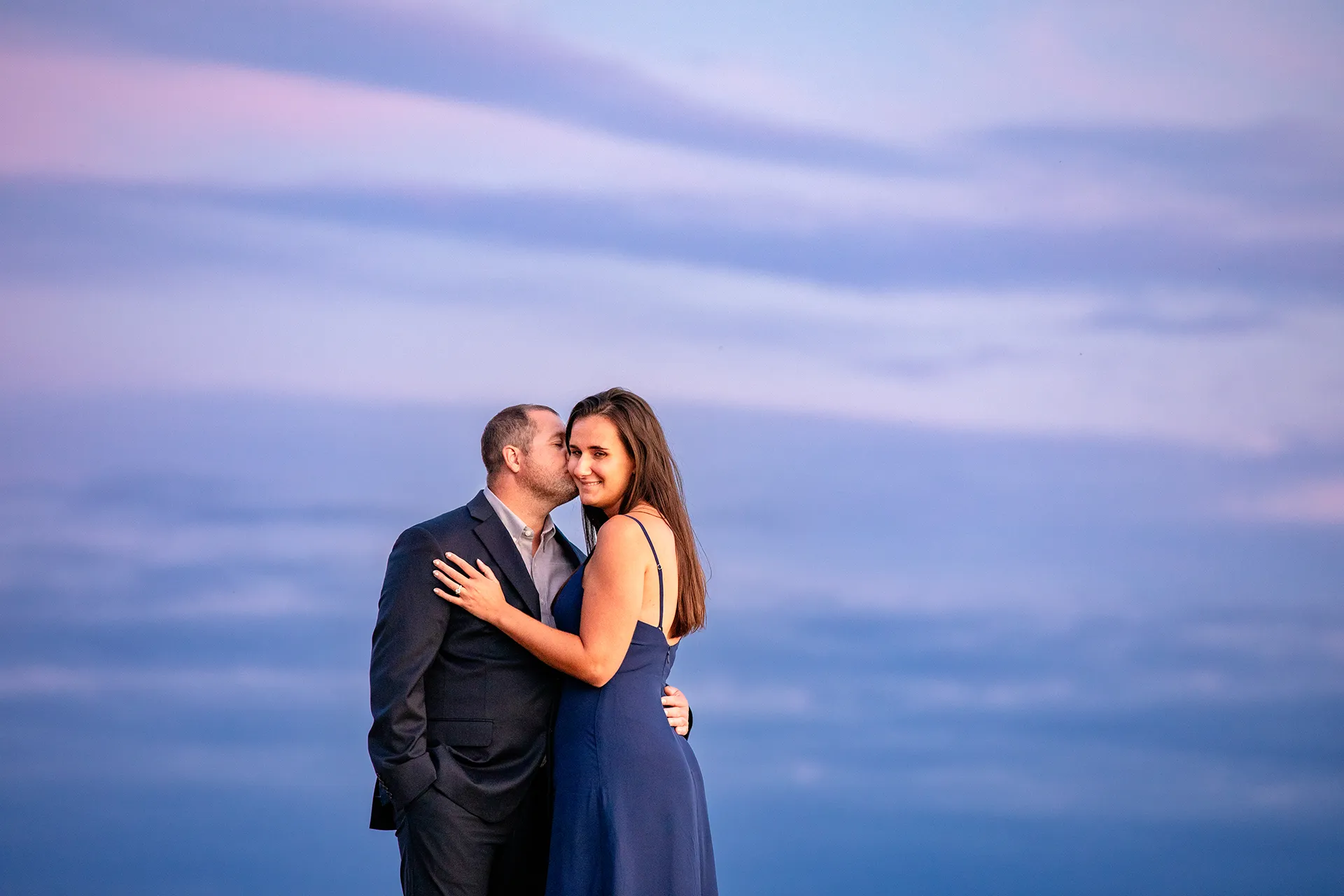 A man kisses a woman on the cheek as the sun sets during an engagement session on Otter Point in Acadia National Park near Bar Harbor, Maine.