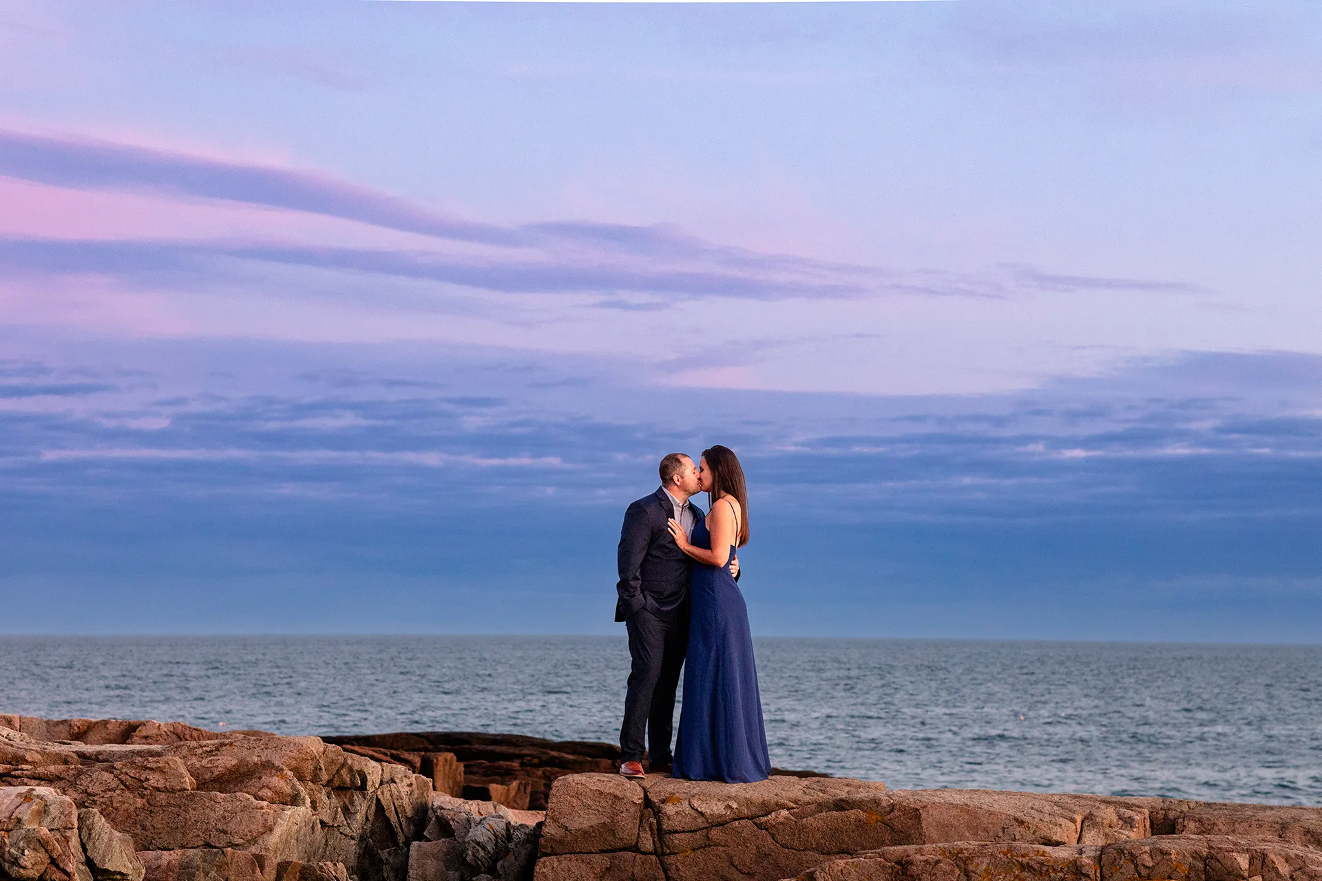 An engaged couple kiss on a cliff during a sunset engagement session on Otter Point in Acadia National Park near Bar Harbor, Maine.
