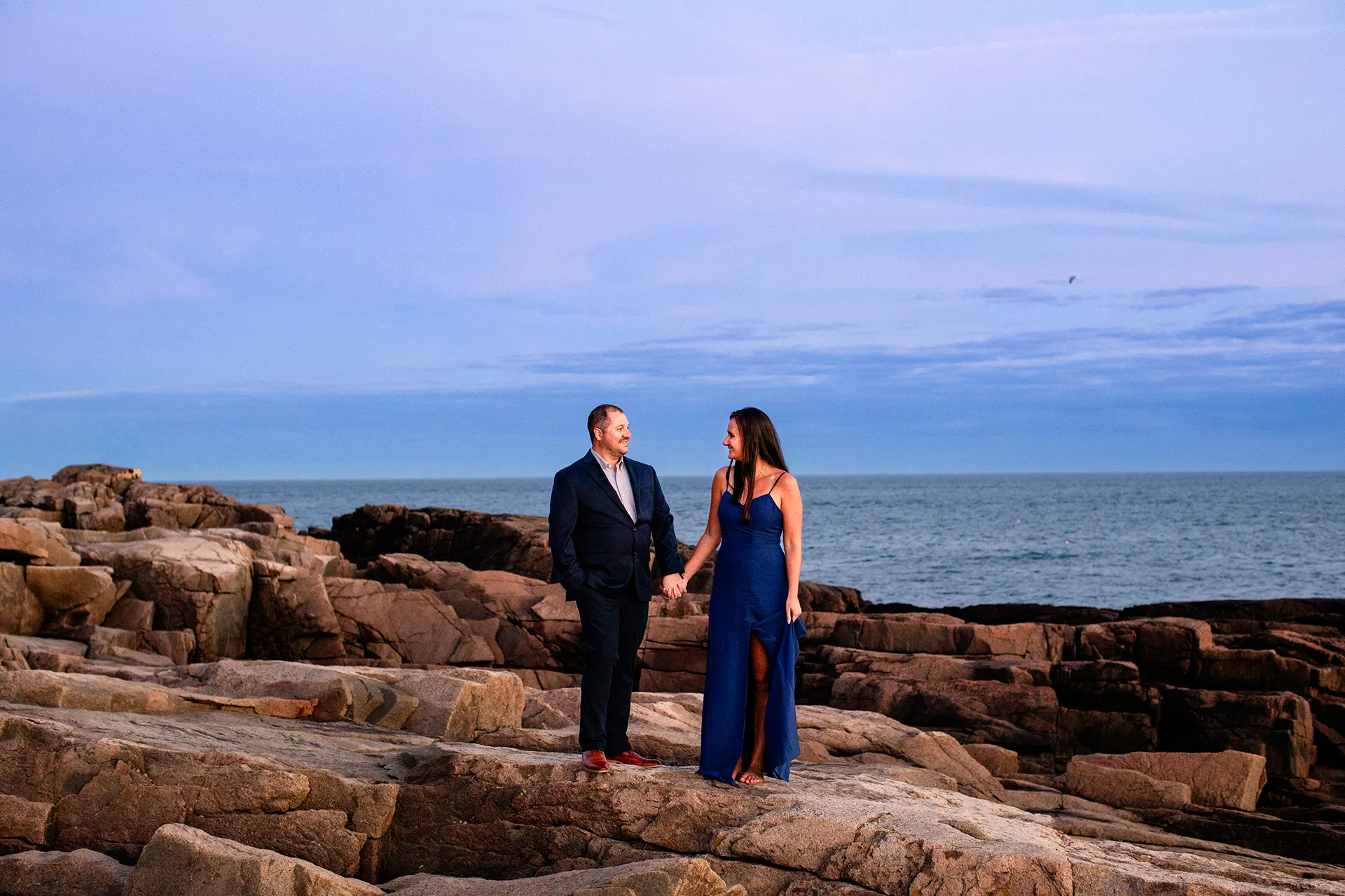 An engaged couple hold hands and smile at each other during an engagement session on Otter Point in Acadia National Park, Maine.