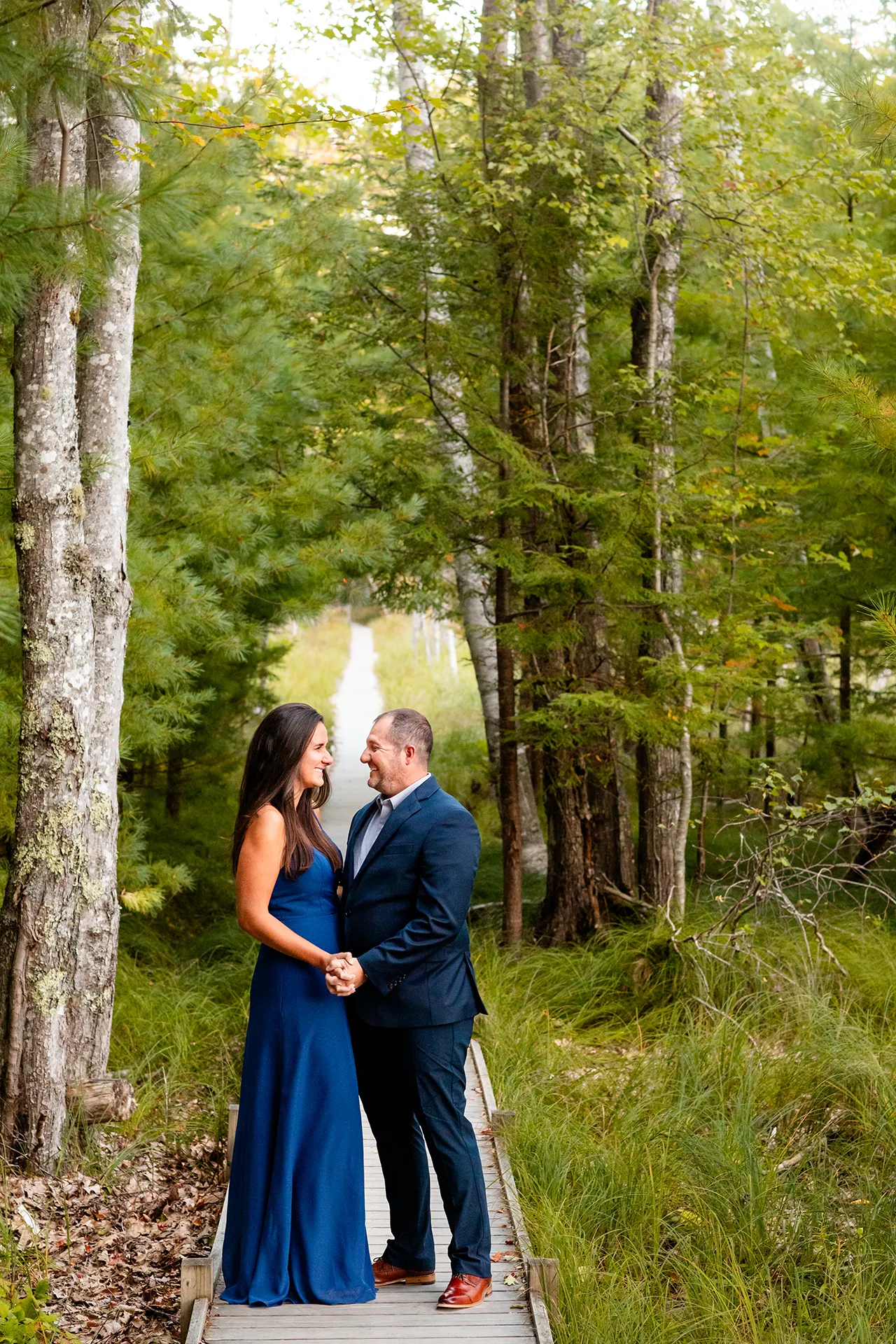 An engaged couple hold hands and smile at each other during a engagement session on the Jesup Path in Acadia National Park, Maine.