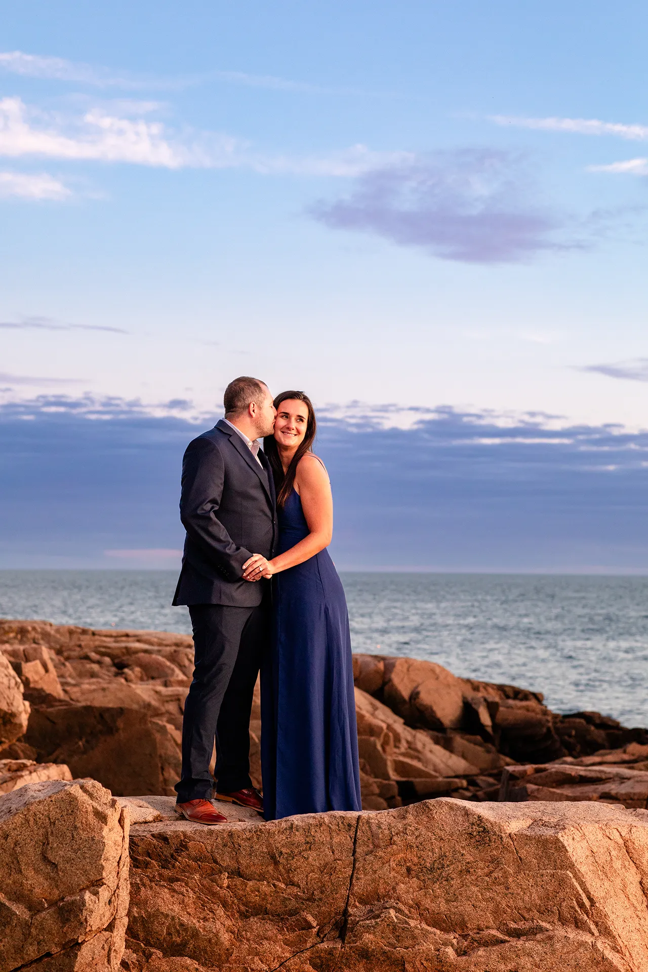 A woman smiles as a man kisses her on the cheek during an engagement session on Otter Point in Acadia National Park near Bar Harbor, Maine.