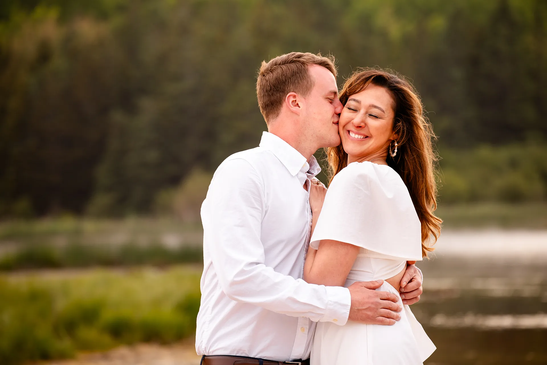 A woman smiles as a man kisses her on the cheek during an engagement session at Sand Beach in Acadia National Park near Bar Harbor, Maine.