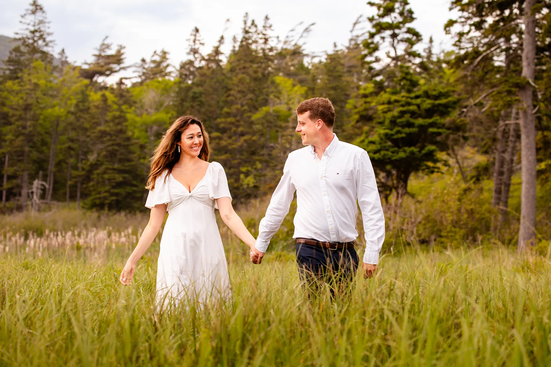 A man and woman hold hands as they walk through a field of grass during an engagement session at Sand Beach in Acadia National Park.