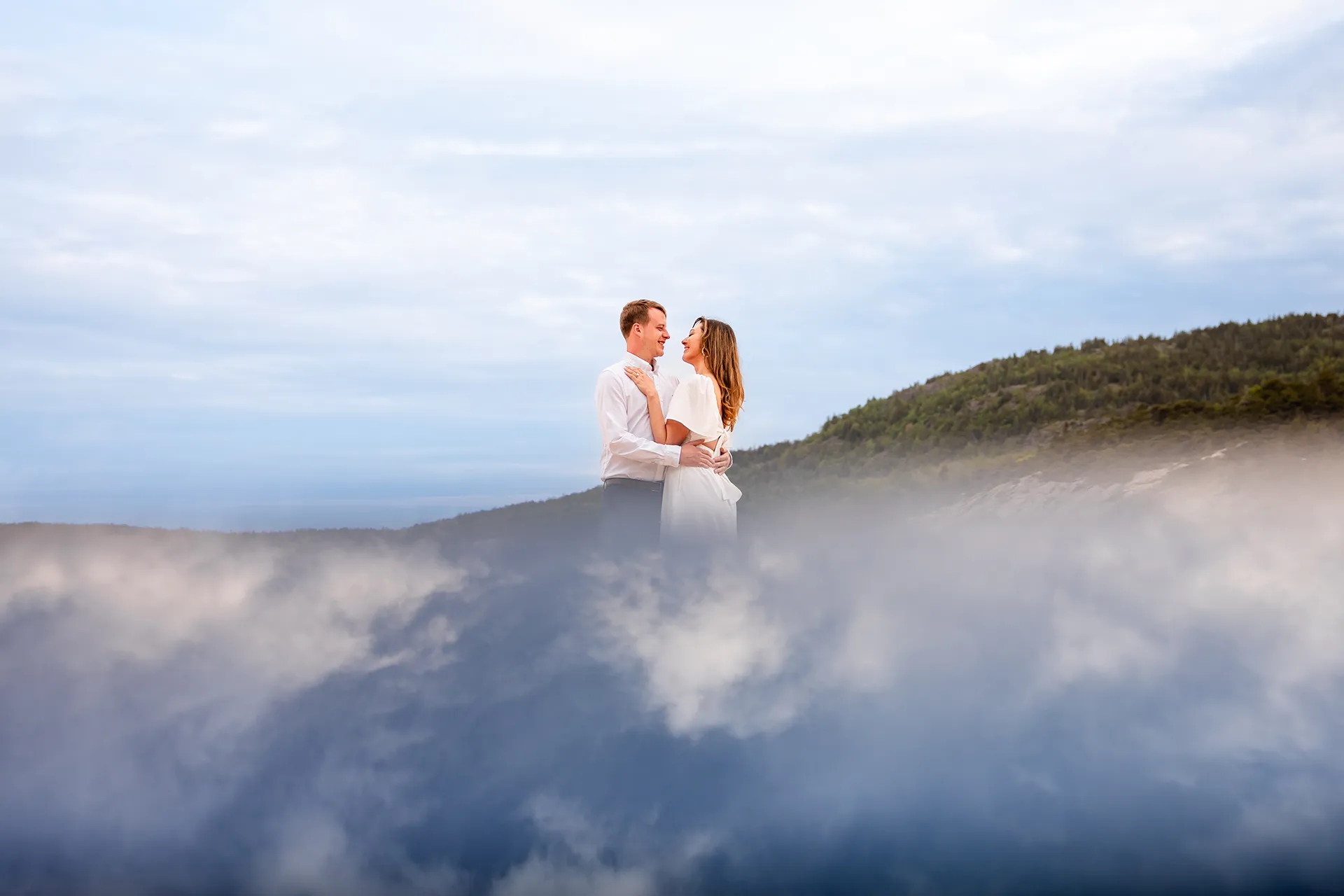 An engaged couple smile at each other while surrounded by blue and white clouds during an engagement session at Sand Beach in Acadia National Park.