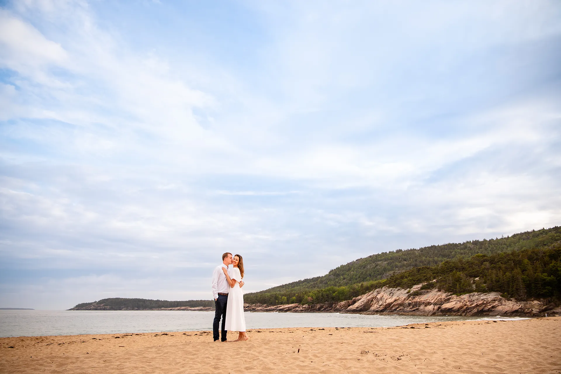 A woman smiles as a man kisses her on the cheek during engagement portraits at Sand Beach in Acadia National Park near Bar Harbor, Maine.