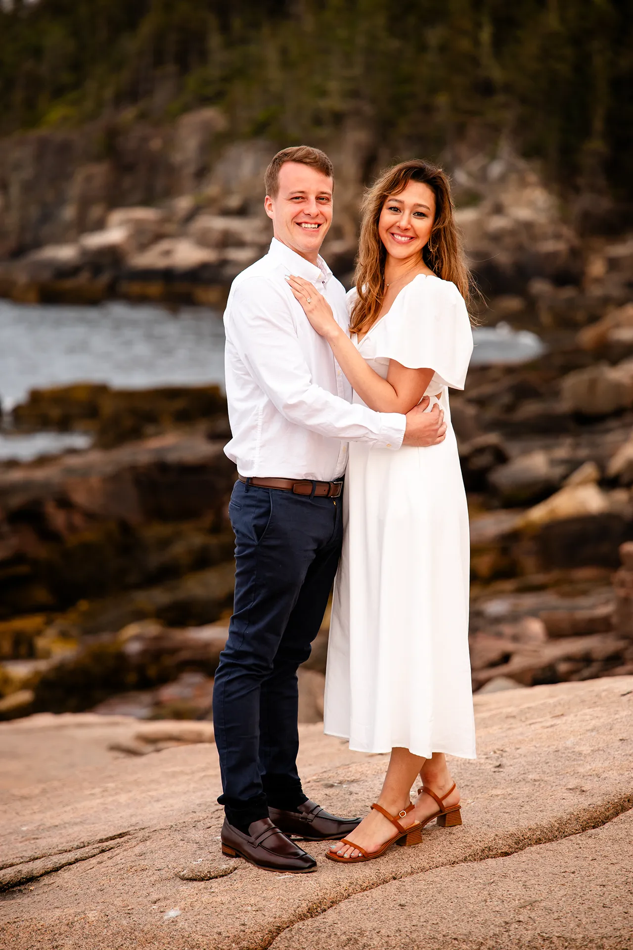 A man and woman smiles and pose during an engagement session near the Ocean Path at Acadia National Park in Bar Harbor, Maine.
