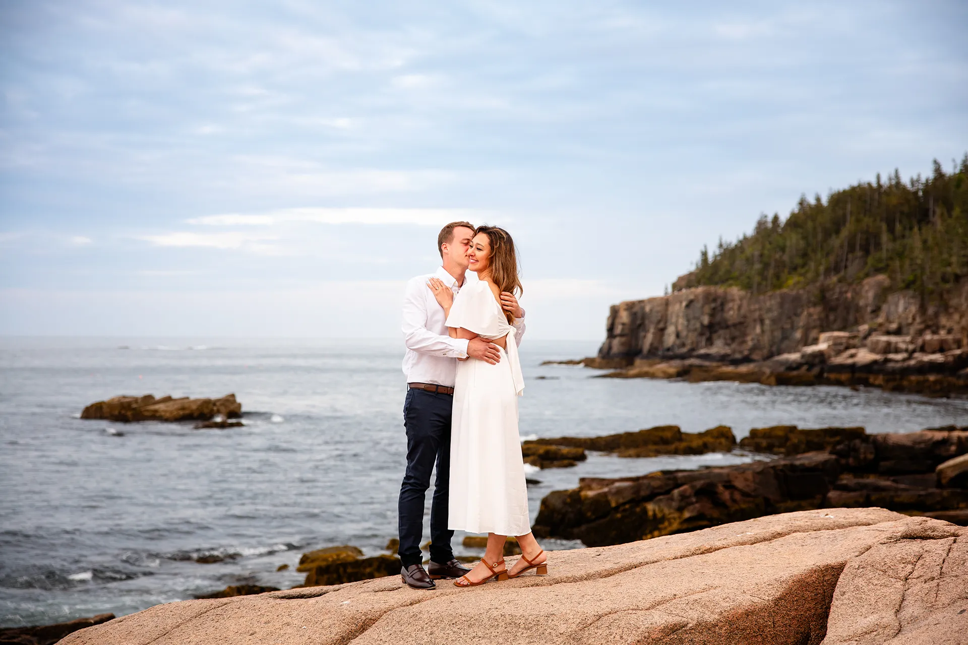 A woman smiling as a man kisses her on the cheek during an engagement session near Otter Cliff in Acadia National Park near Bar Harbor, Maine.