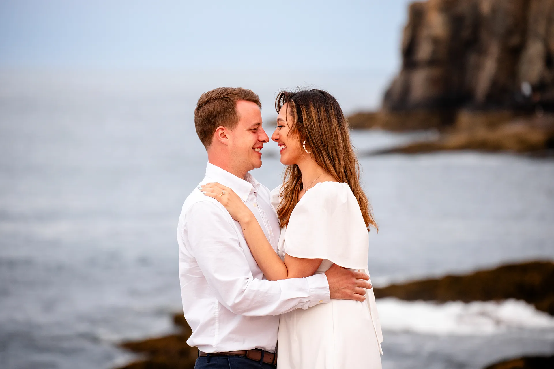A man and woman laugh as they look at each other during a coastal engagement session near Otter Cliff in Acadia National Park, Maine.