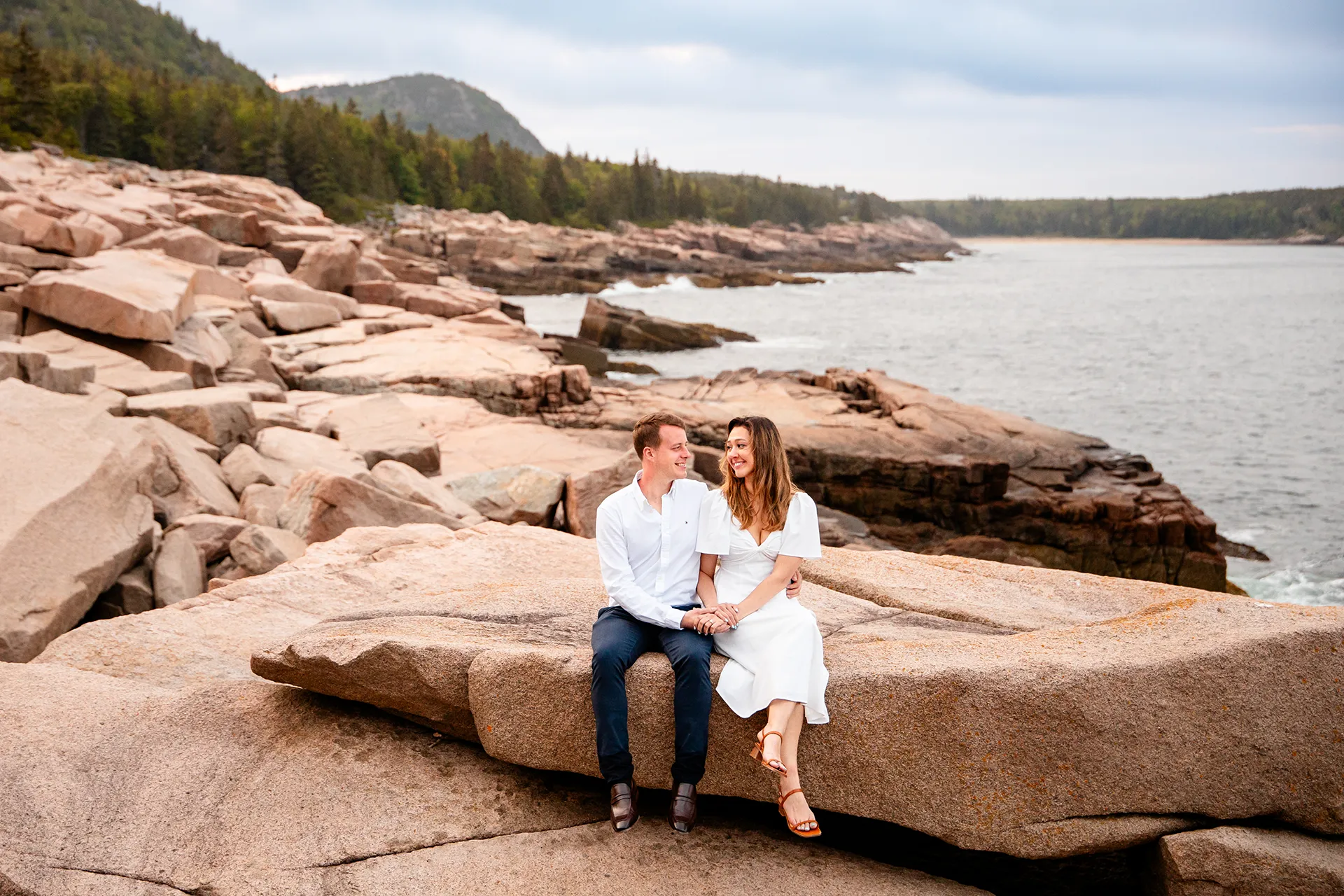 An engaged couple sit on a rock and hold hands during an engagement session near Beehive Mountain in Acadia National Park, Maine.