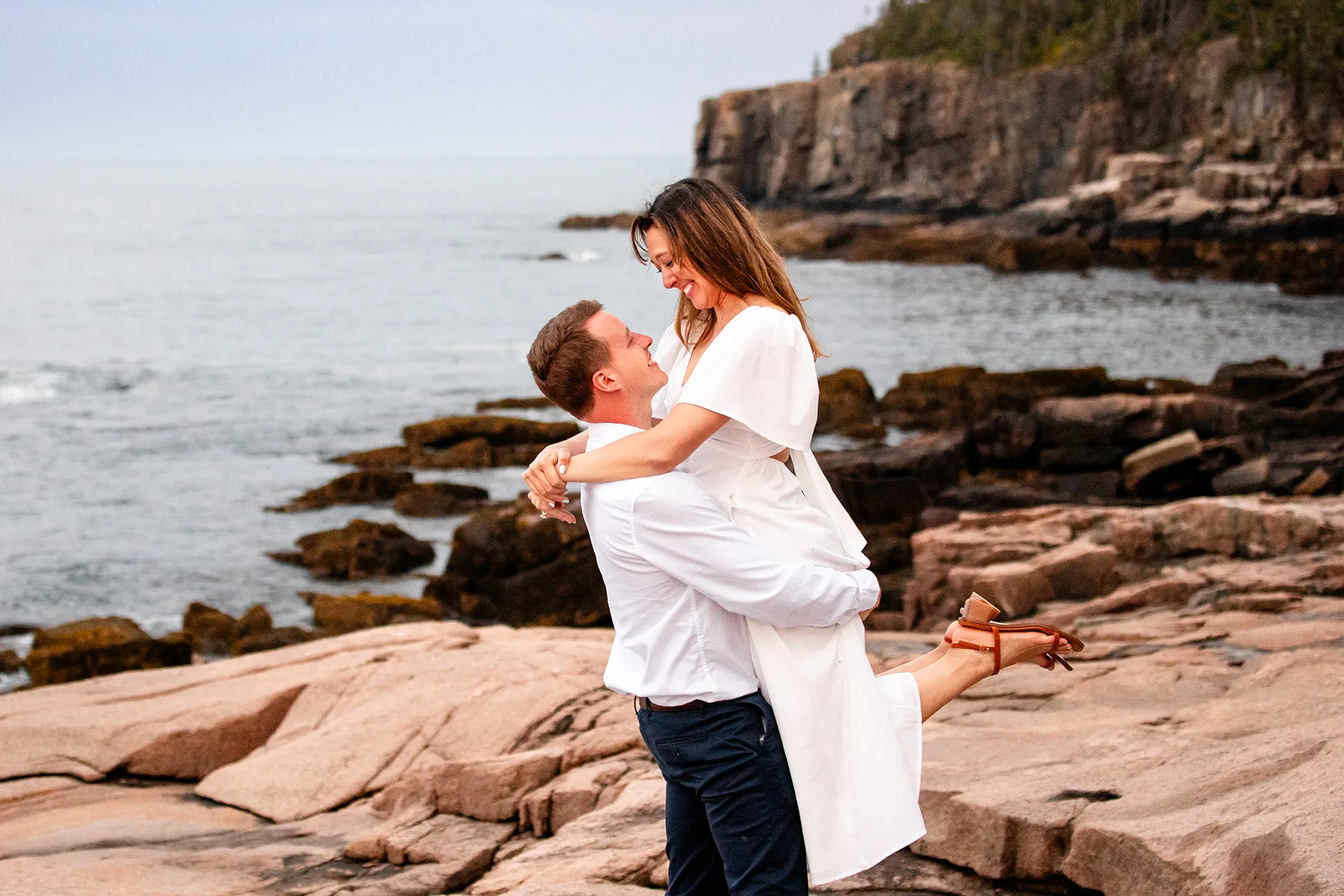 A man lifts a woman during a coastal engagement session near the Ocean Path in Acadia National Park near Bar Harbor, Maine.