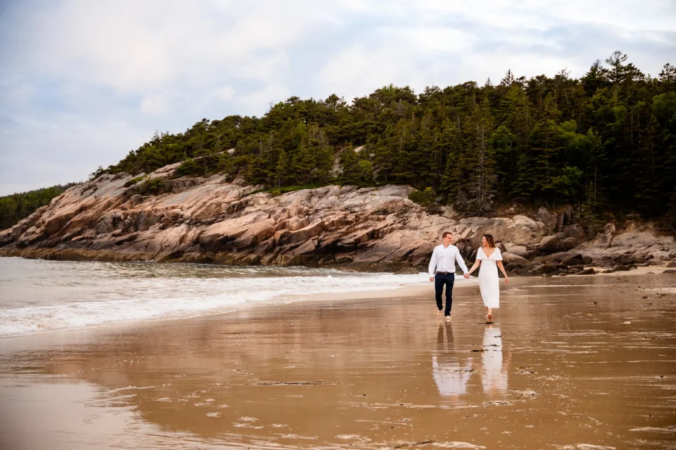 A man and woman hold hands and walk on Sand Beach during engagement session in Acadia National Park near Bar Harbor, Maine.