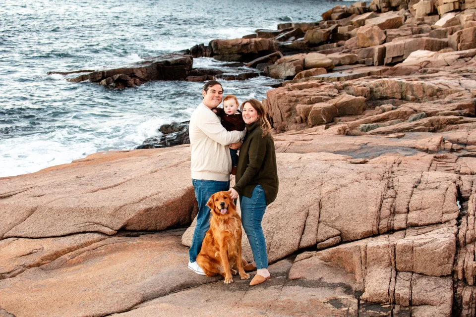 A dad holds his son as a mom smile and pats a golden retriever during a family portrait session near Otter Cliff in Acadia National Park.