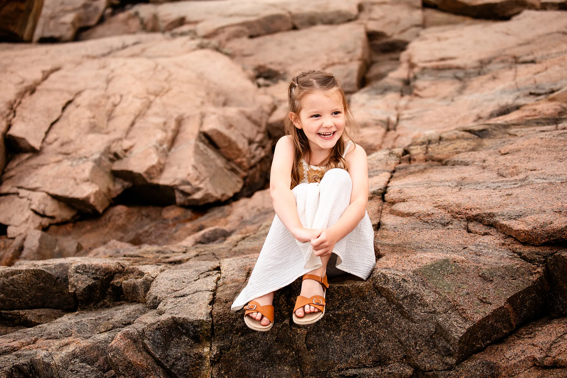 A girl sits on a rock and laughers during family portraits near the Ocean Path at Acadia National Park in Bar Harbor, Maine.