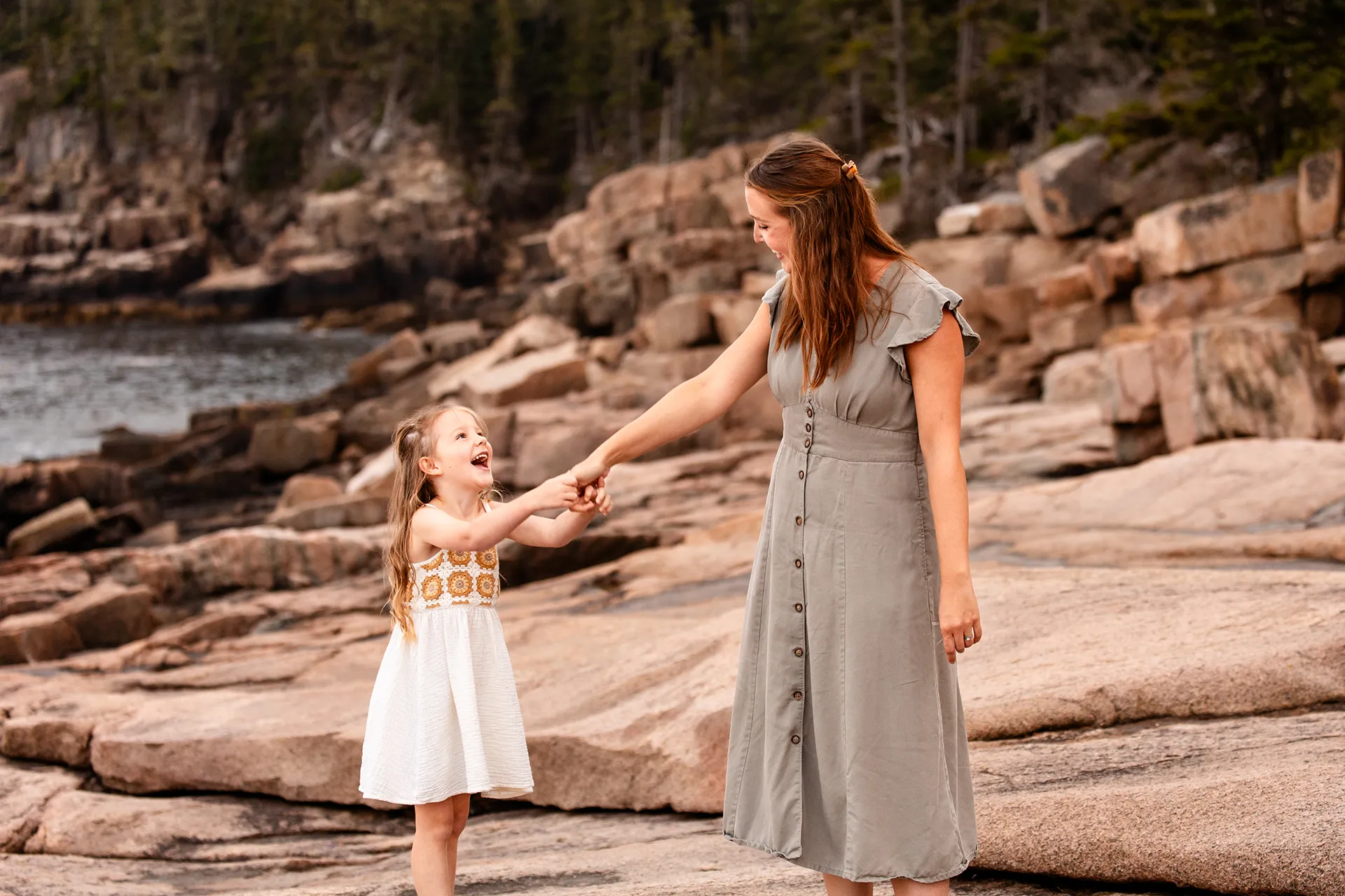 A girl holds her mom's hands and laughs near Otter Cliff during family portraits at Acadia National Park in Bar Harbor, Maine.