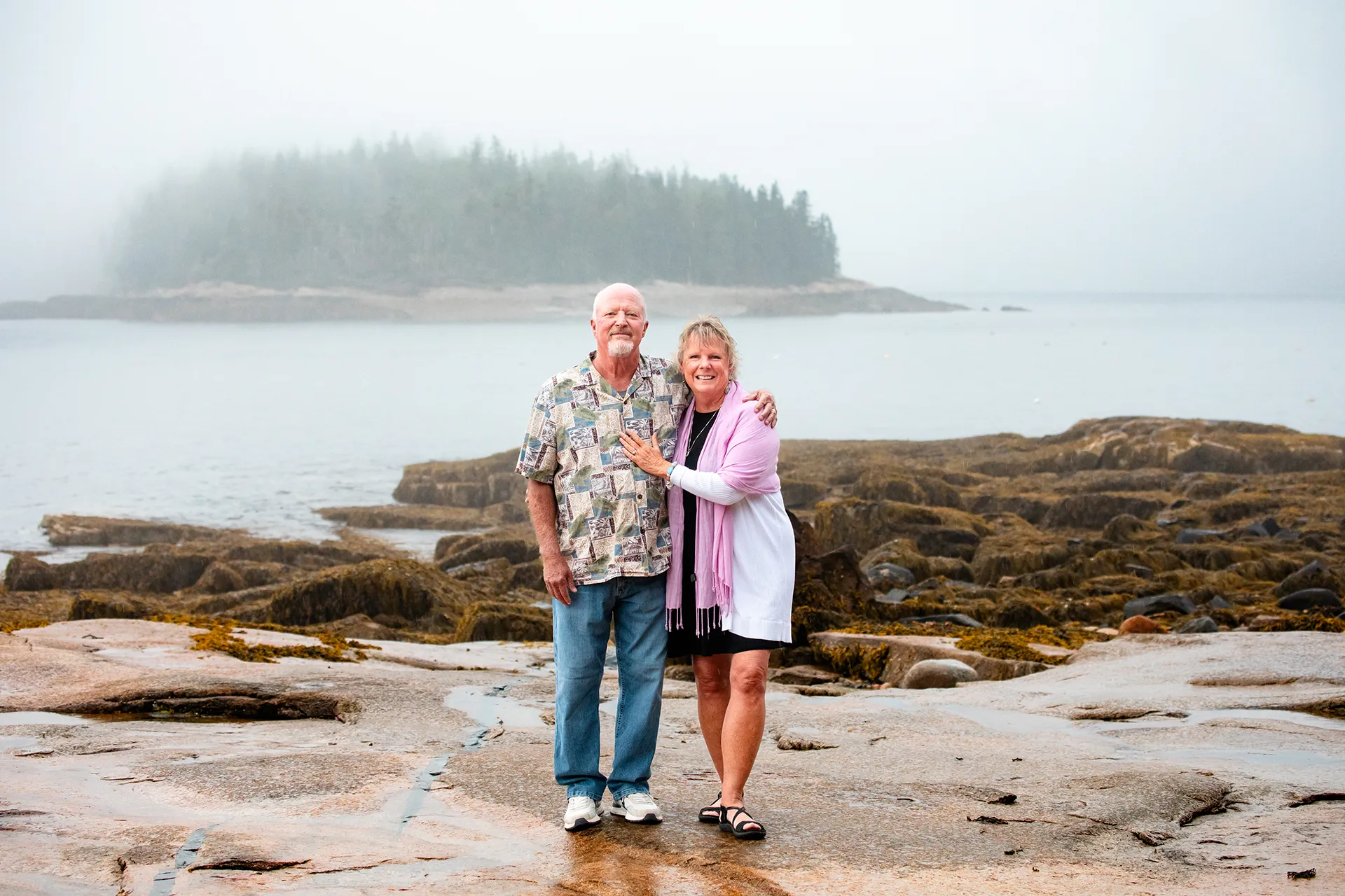 A man and women smile and pose in front of a foggy harbor at Grindstone Point in Winter Harbor, Maine.