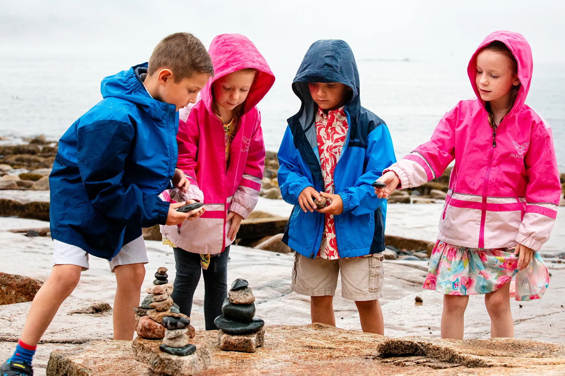 Children wearing rain jackets stack rocks during a family portrait session Grindstone Point in Winter Harbor, Maine.
