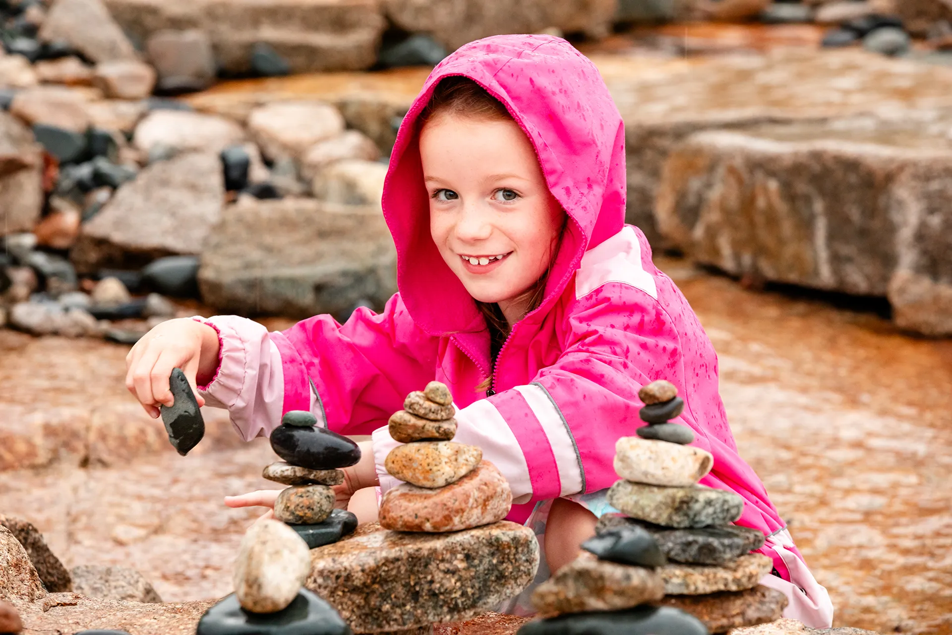 A girl smiles as she stacks rocks in the rain during a family portrait session Grindstone Point in Winter Harbor, Maine.
