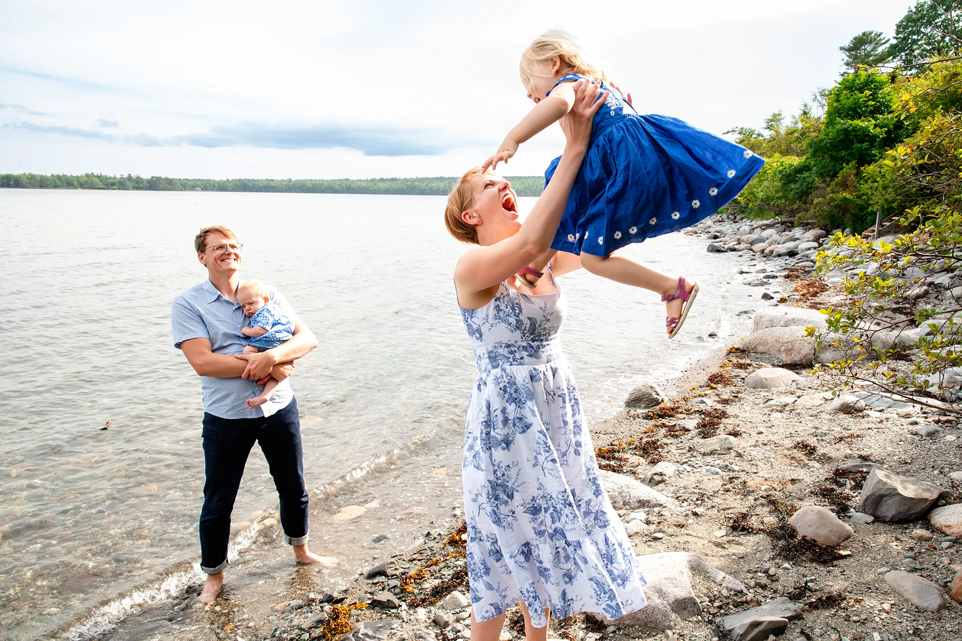 maine family photography moldawer mandsager 09 A mom laughs as she lifts a young girl in the air as a dad watches during a family portrait session in Lamoine, Maine.