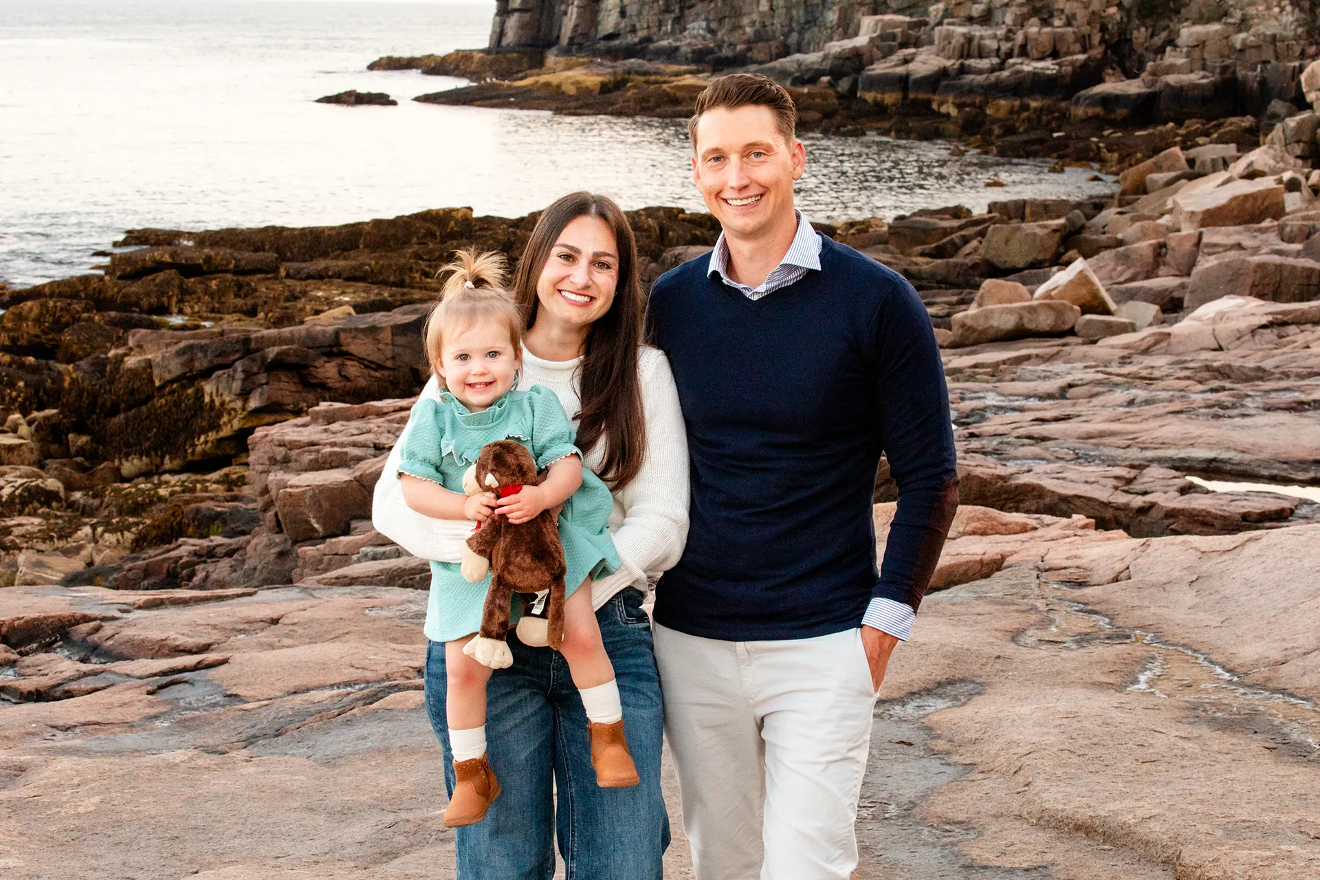 maine family photography myer 14 A mom smiles as she holds her daughter and a dad smiles and poses during a family portrait session near the Otter Cliff in Acadia National Park.