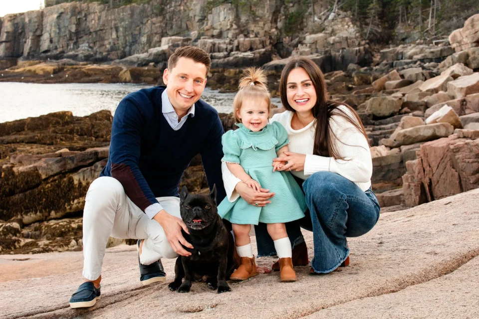 A mom and dad smiles and poses with their daughter and black dog during a family portrait session near the Otter Cliff in Acadia National Park.