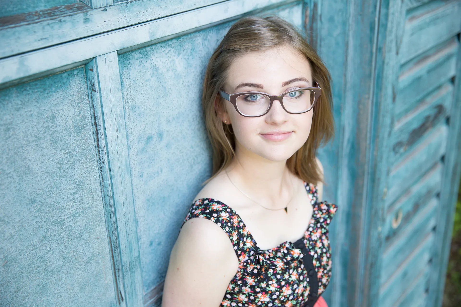 maine portrait photography emma 02 A girl smiles and poses against a green shed during a senior portrait session in Littlefield Ornamental Garden at the University of Maine in Orono.