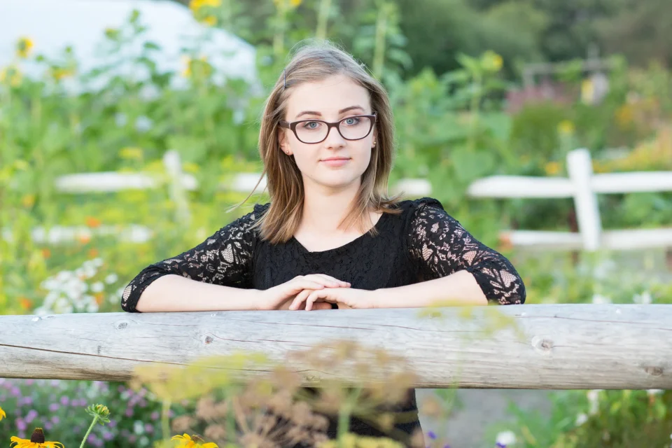 A girl rests her hands on a fence while posing in a garden during a senior portrait session at Rogers Farmer near the University of Maine in Orono.