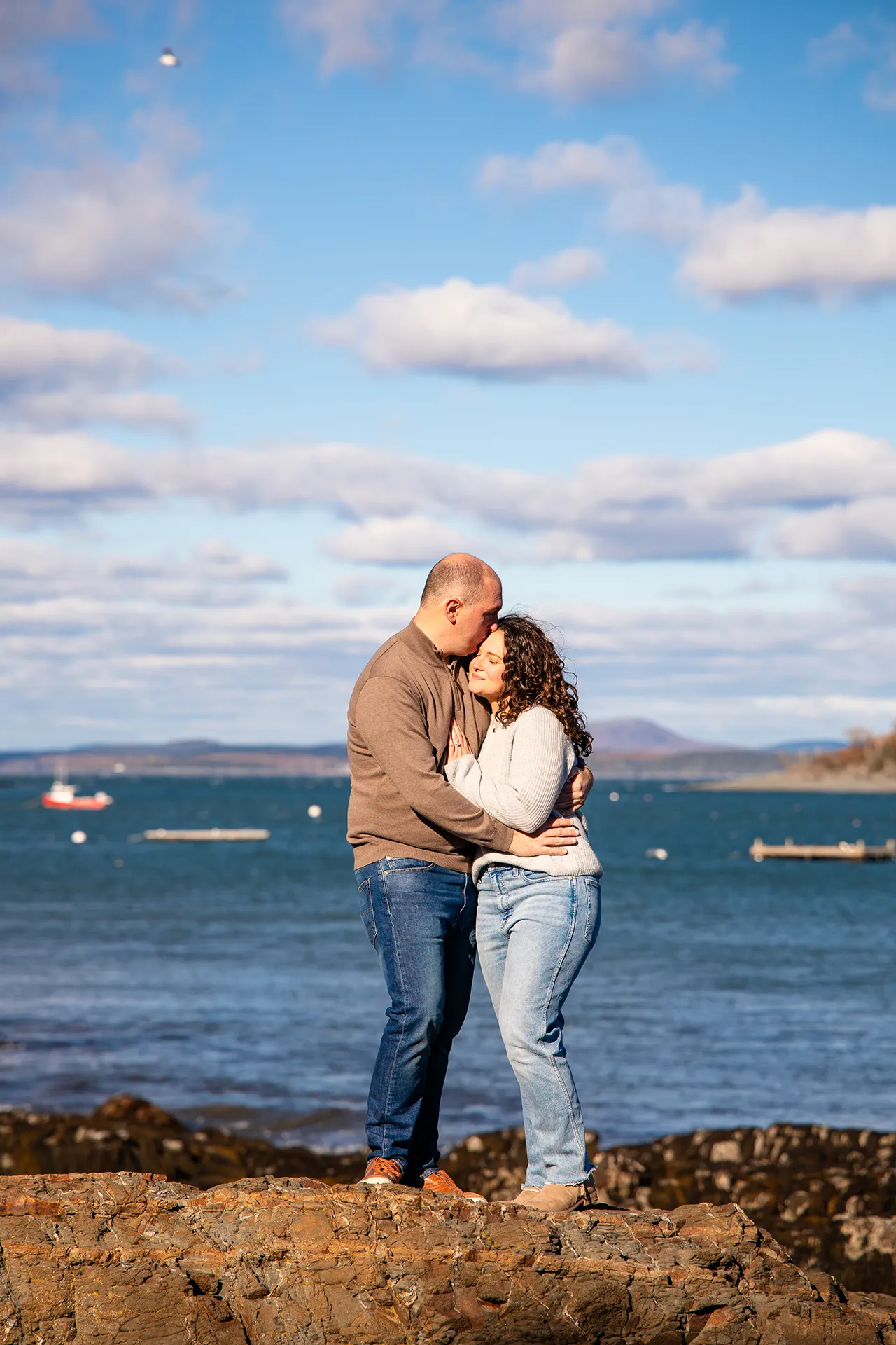A man kisses a woman on the forehead while they stand in front of Frenchmans Bay during engagement portrait at the Bar Harbor Inn and Spa.