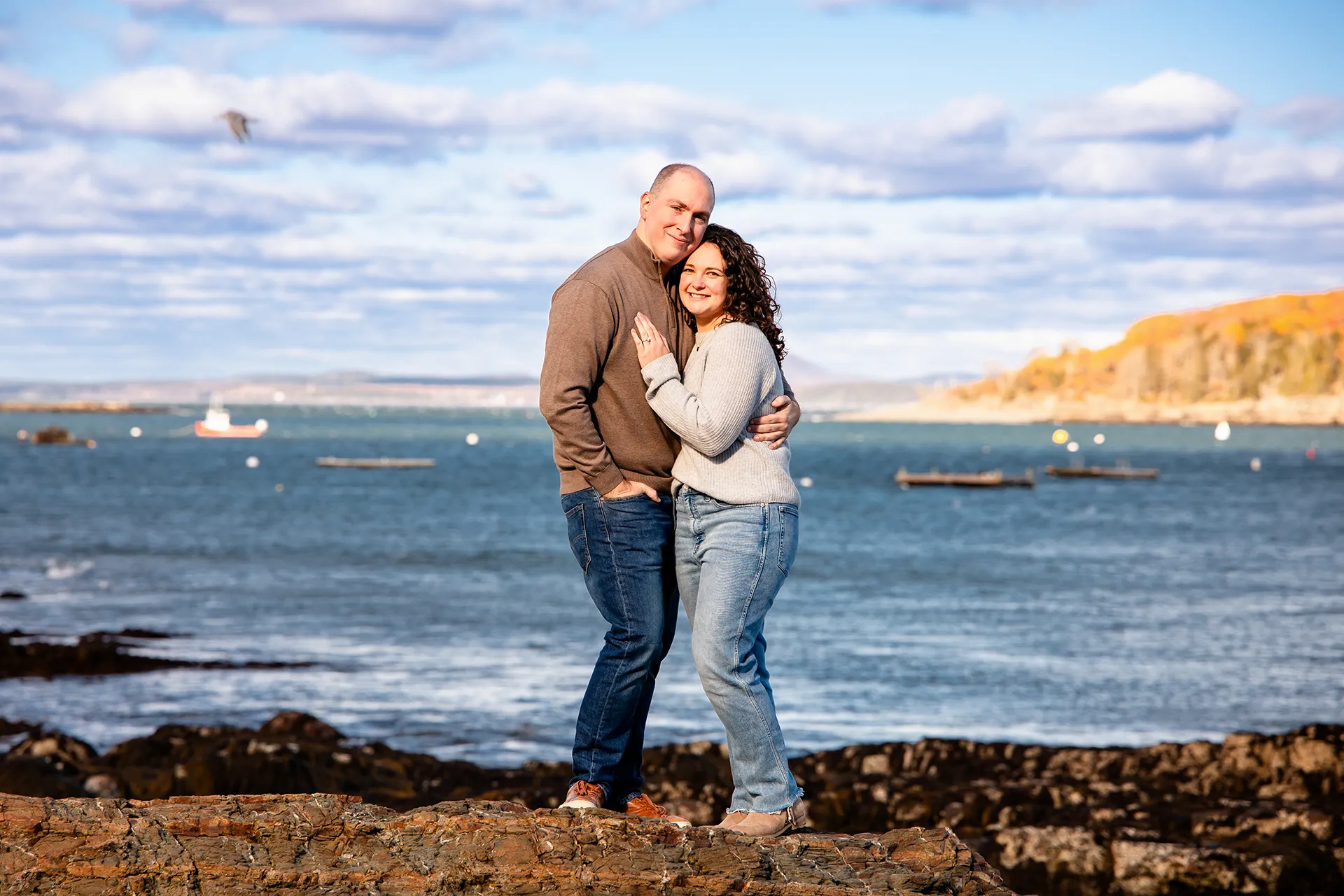 A man and woman smile and pose in front of Frenchmans Bay during engagement portrait at the Bar Harbor Inn and Spa in Maine.